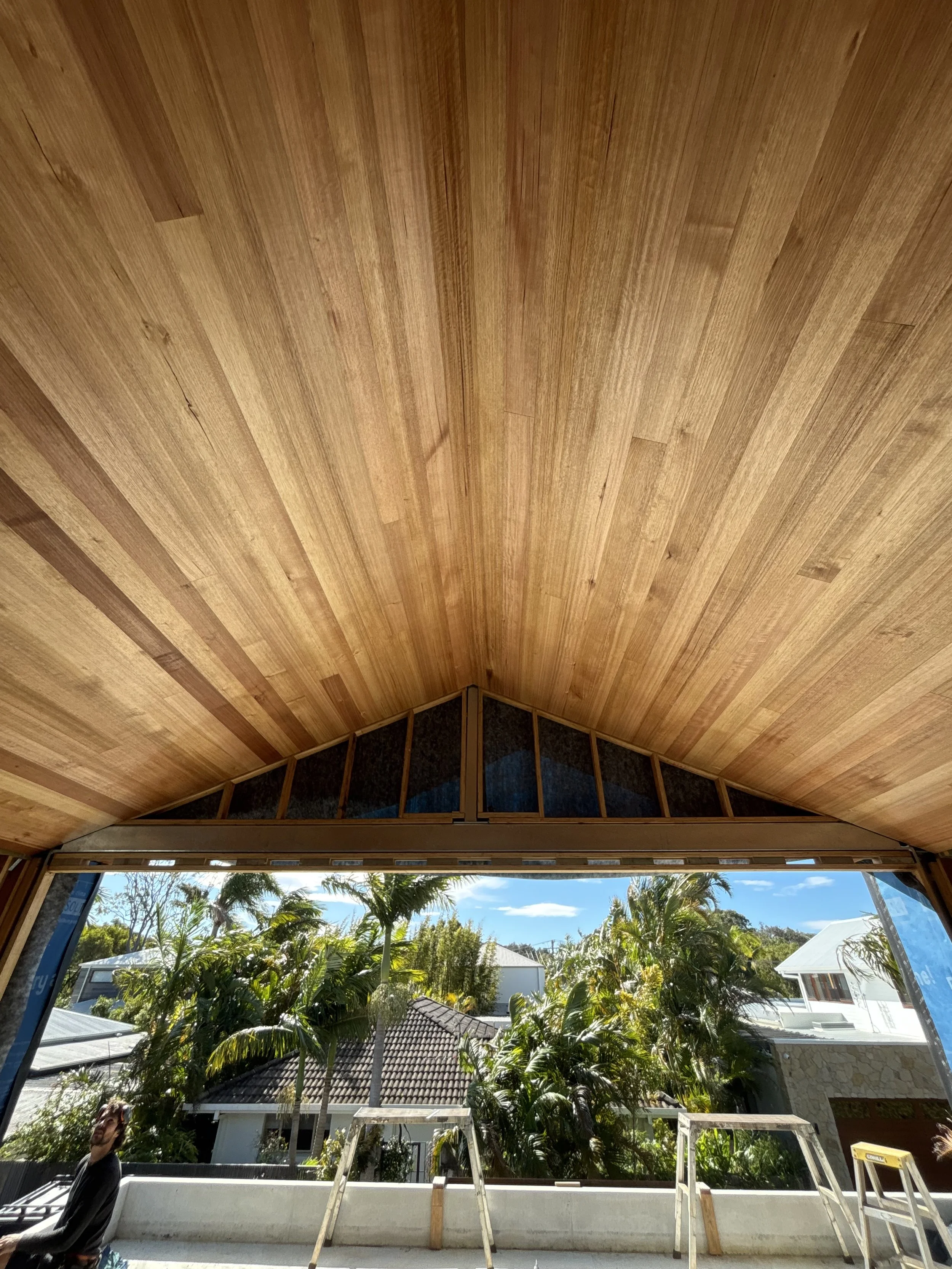 Interior view of a room under construction with a wooden ceiling and a large window showing trees and houses outside.