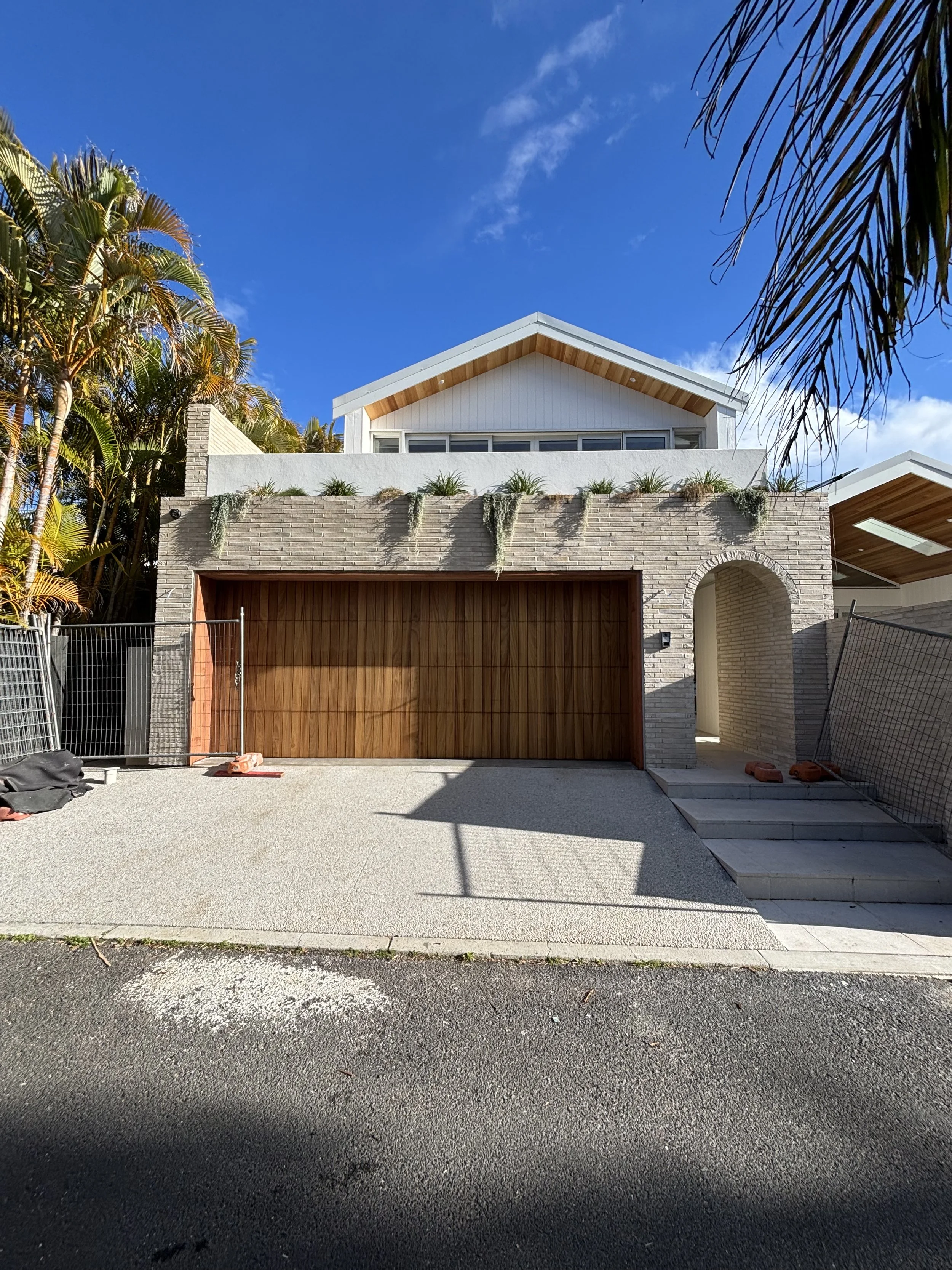 A modern house with a driveway, surrounded by palm trees, under a clear blue sky with some clouds.