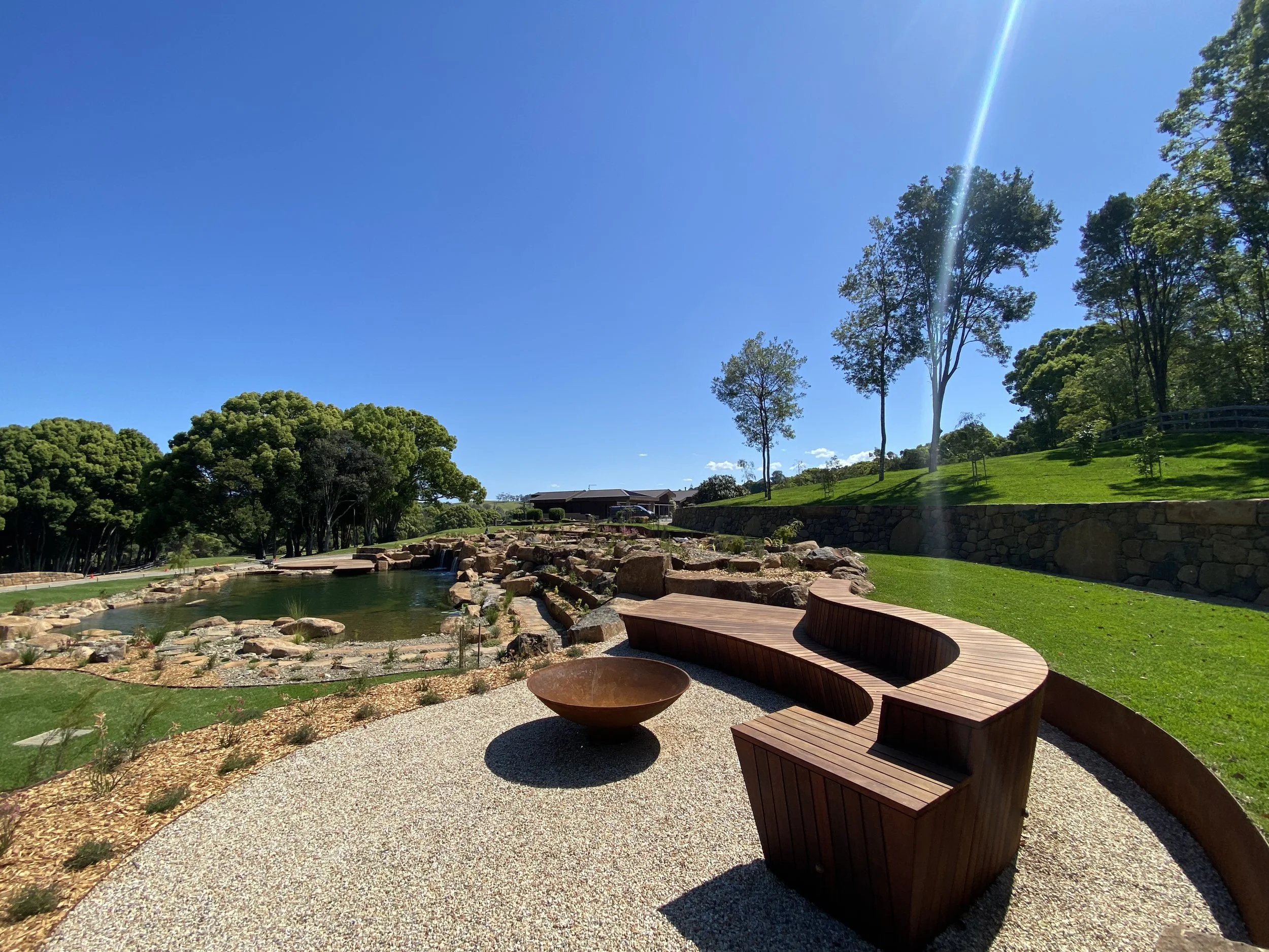 A sunny park with a small pond, stone border, a curved wooden bench, and lush green trees under a clear blue sky.