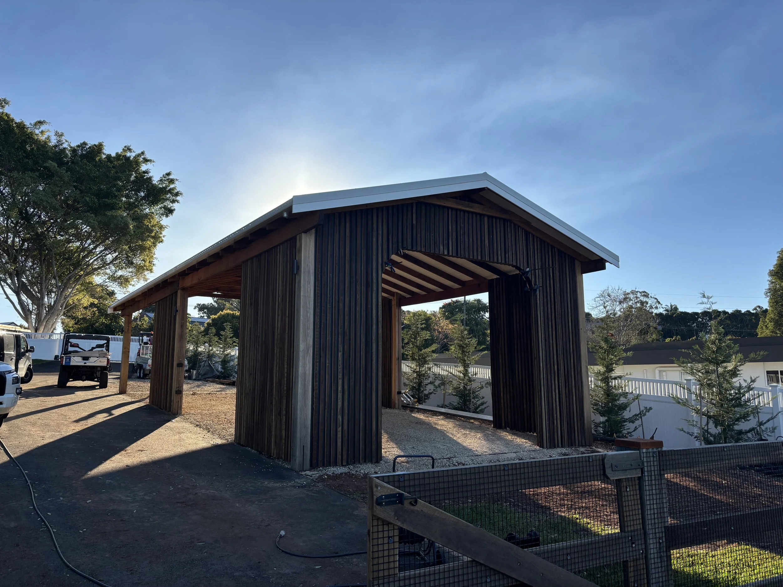 A wooden structure under construction with a metal roof, surrounded by trees and a parking area with parked vehicles, and a white fence in the background.