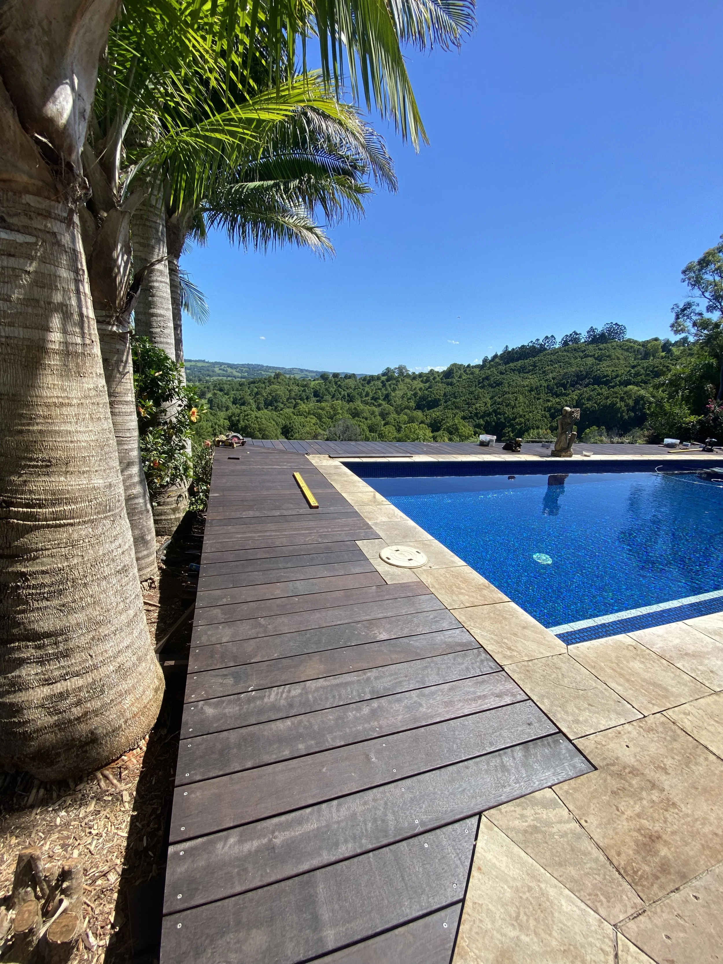 View of a backyard with a rectangular swimming pool, wooden deck, palm trees, a small statue, and lush green hills in the background under a clear blue sky.