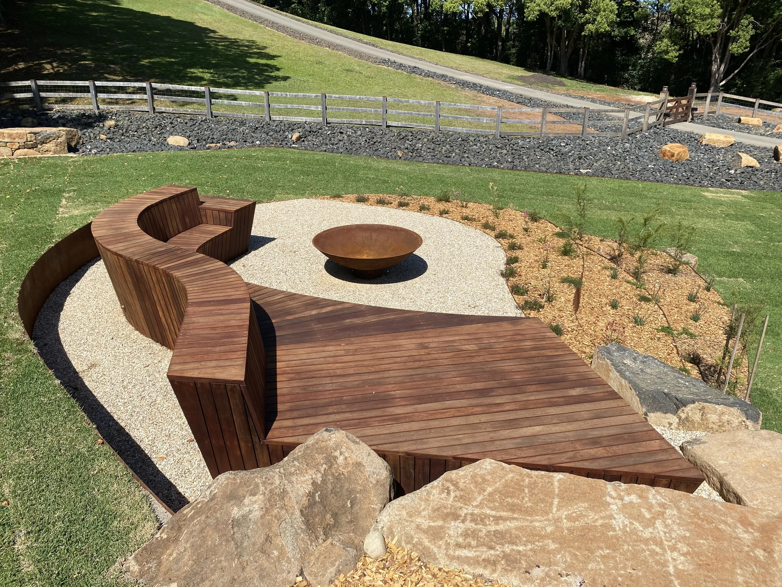 A landscaped outdoor area with a curved wooden bench, a fire pit, and a garden bed with small plants. There is a wooden fence, stone path, and green grass in the background.