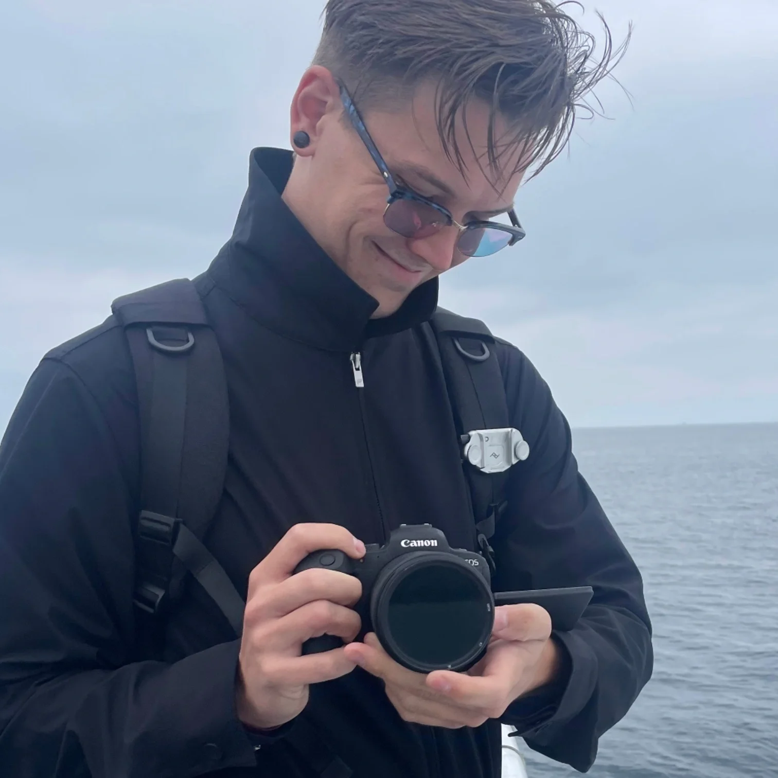 A young man with short hair and sunglasses, wearing a black jacket and carrying a backpack, is holding a Canon camera and looking down, standing outdoors near a body of water under a cloudy sky.