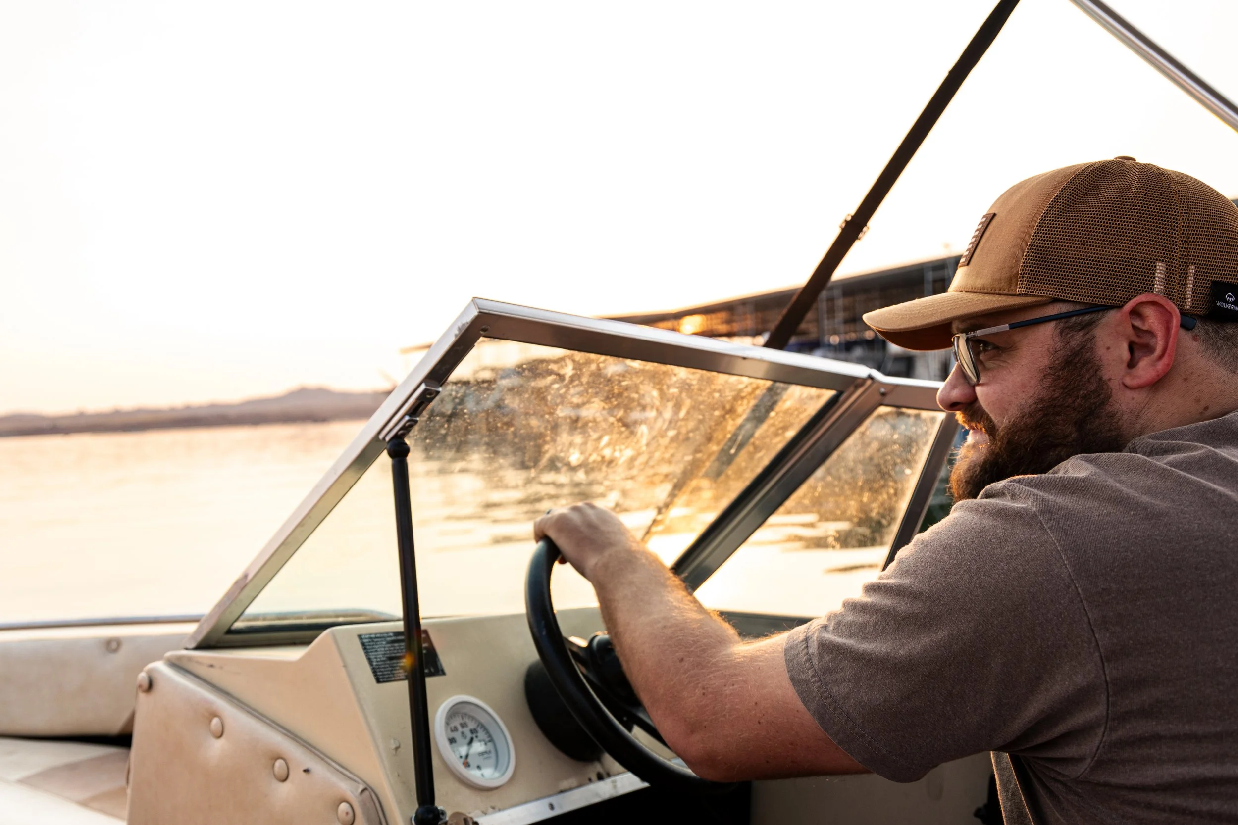A man with a beard and glasses driving a boat on a river during sunset, wearing a tan cap and gray t-shirt.