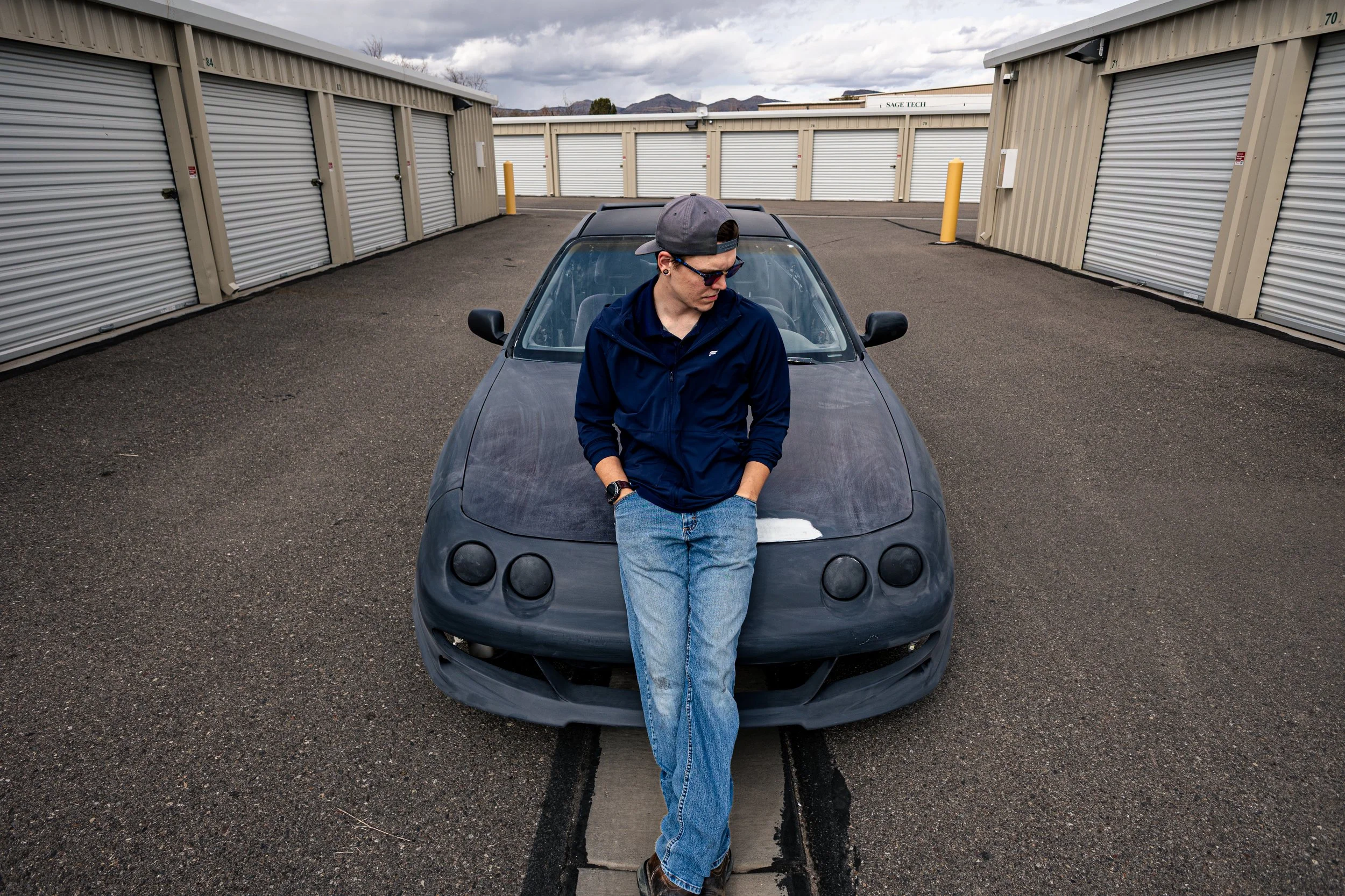 A young man wearing a cap and sunglasses leaning against a black sports car parked in a storage facility lot with storage units in the background.