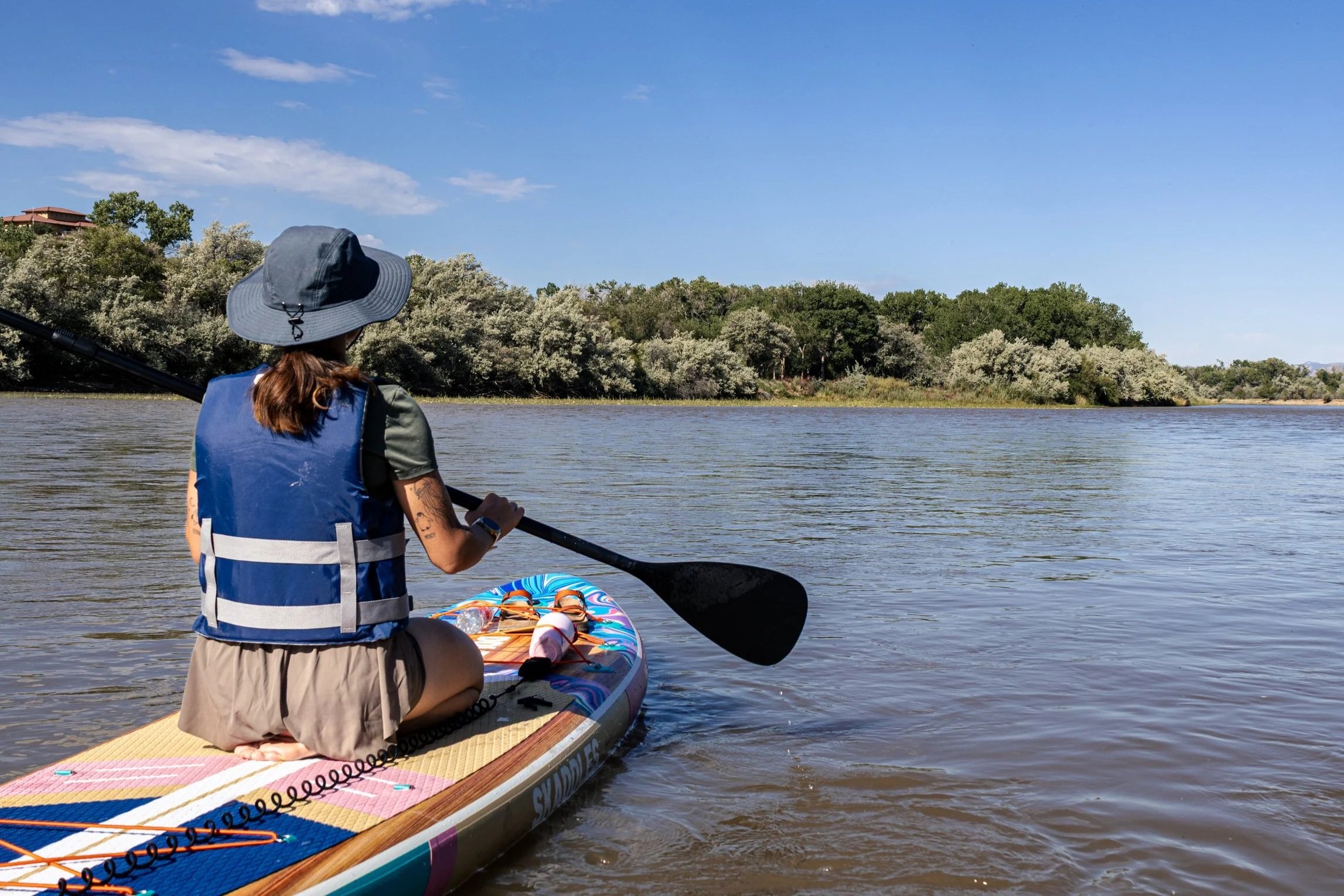 Person wearing a wide-brimmed hat and life jacket sitting on a paddleboard on a river, holding a paddle, with trees and a house in the background under a blue sky.