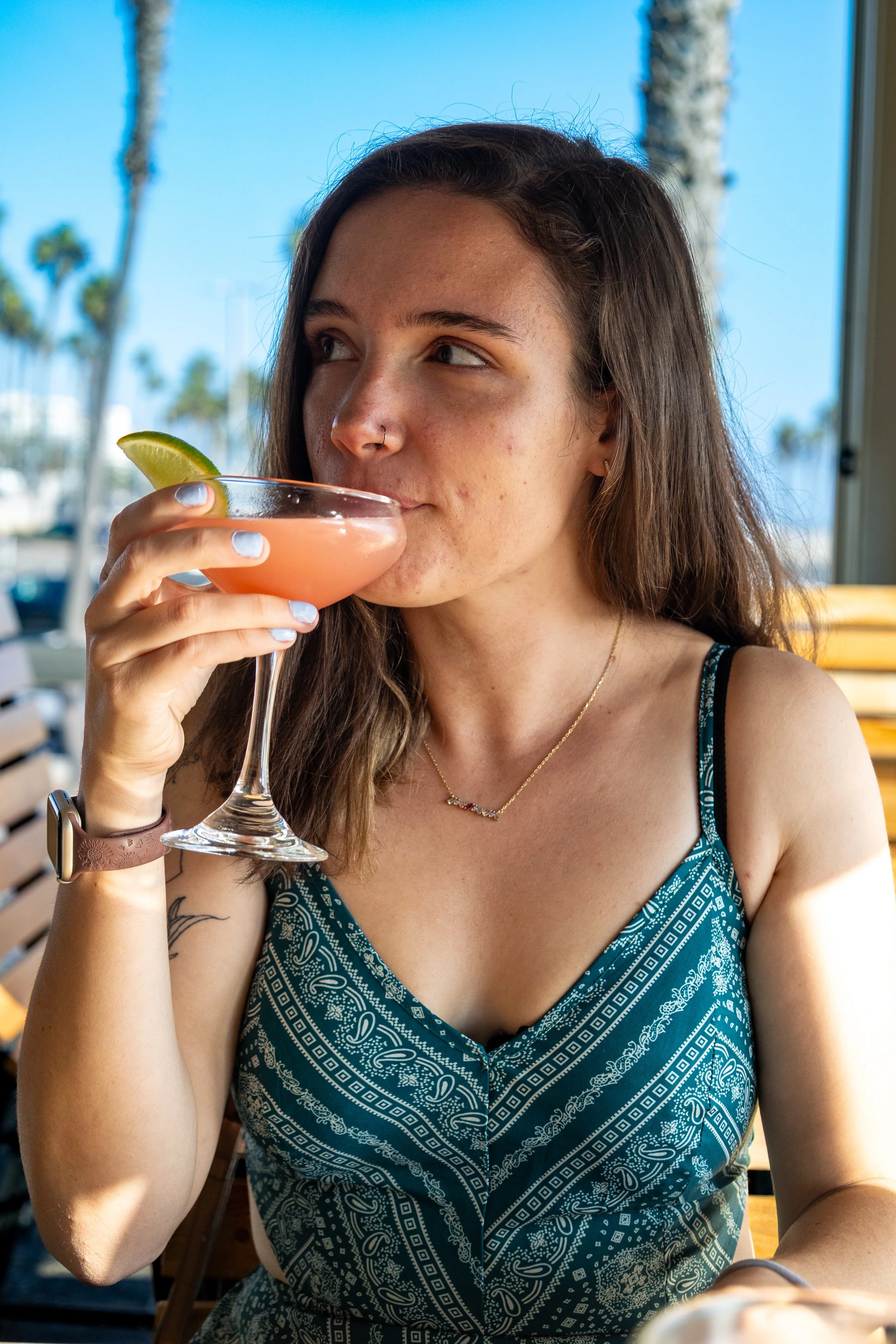 A young woman with long brown hair, wearing a blue sleeveless dress with a paisley pattern, is sipping a pink cocktail with a lime wedge garnish, sitting outdoors on a sunny day with palm trees and blue sky in the background.