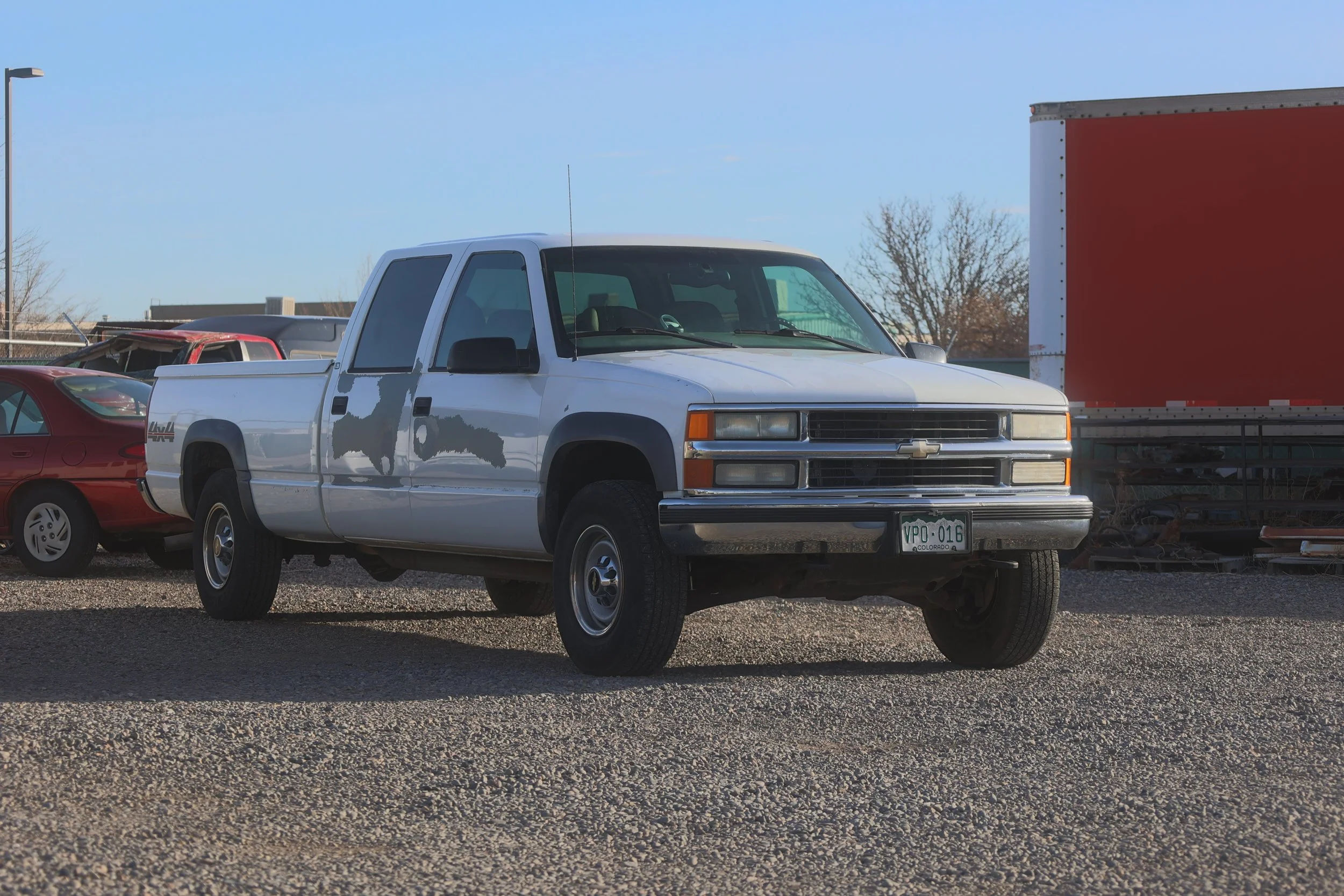 A white Chevrolet pickup truck parked on gravel with an orange-red trailer behind it. Several other vehicles are visible nearby, including a red car and a damaged red vehicle. The sky is clear and blue, with a few trees in the background.