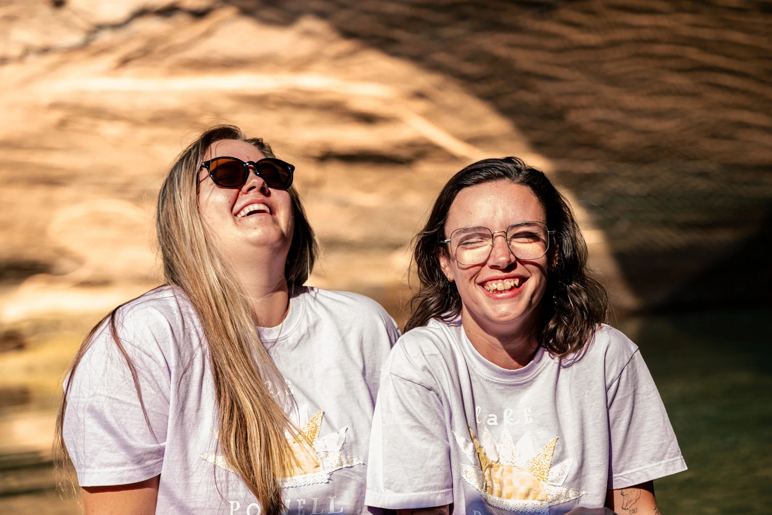 Two women with long hair smiling and wearing glasses, dressed in white T-shirts, standing outdoors with rocky scenery in the background.
