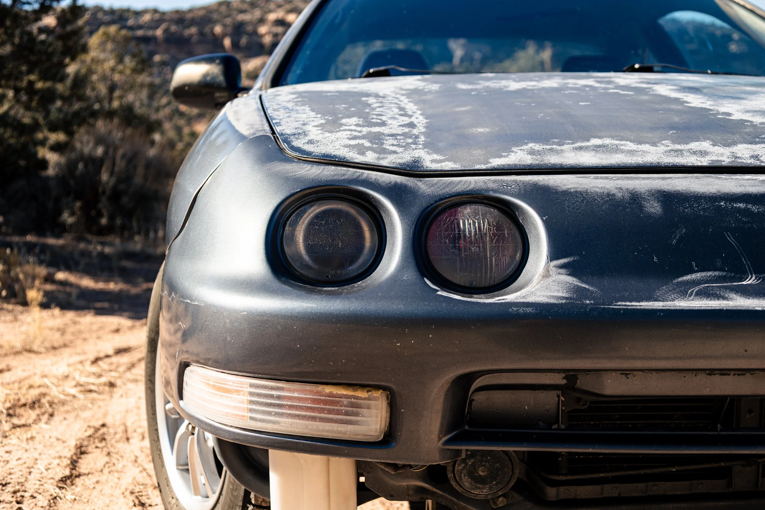 Close-up of the front of a silver car with a dusty hood and round headlights, parked outdoors on a dirt surface with blurred trees in the background.