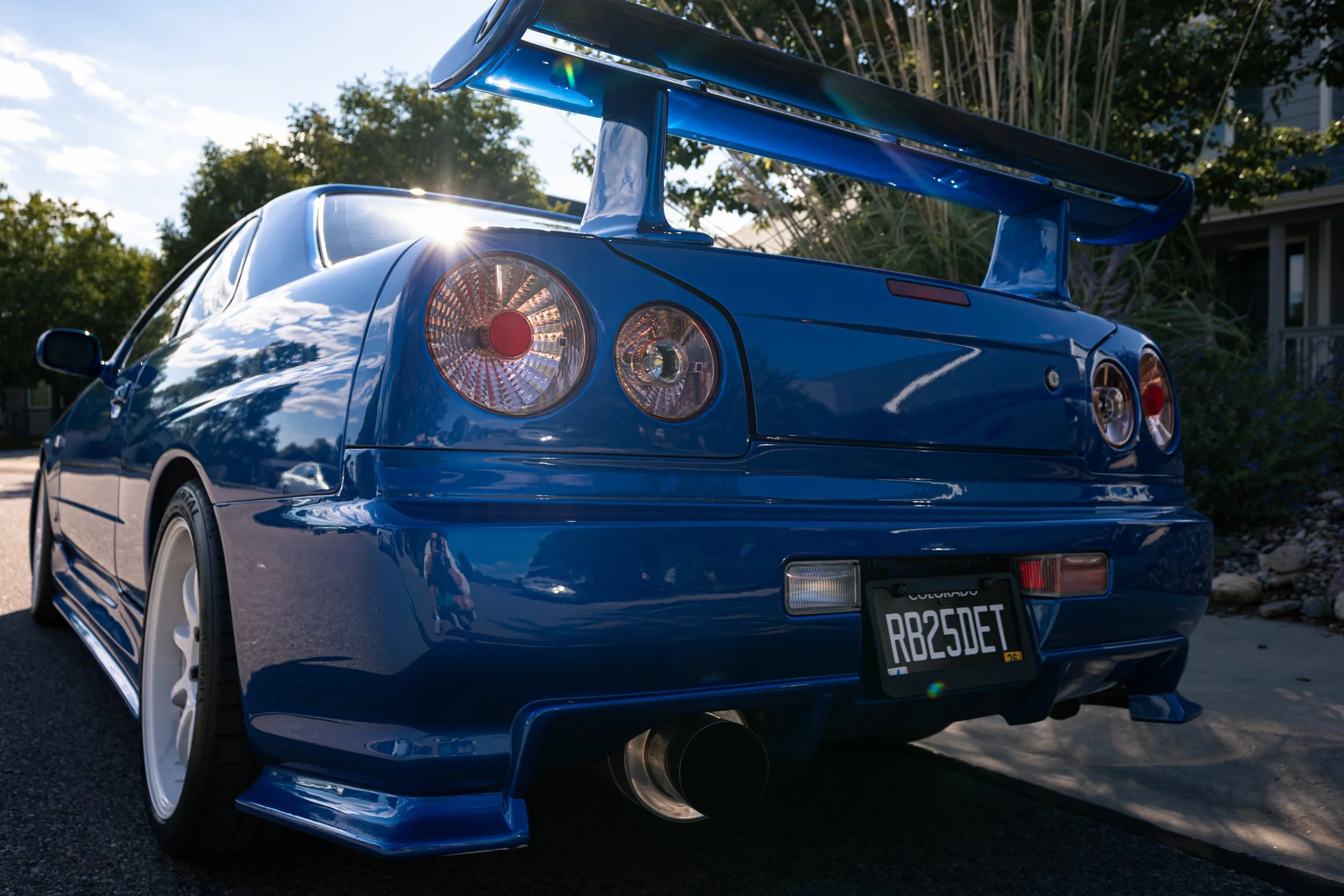A blue sports car with a large rear spoiler, parked outdoors during daylight, with trees and a house in the background.