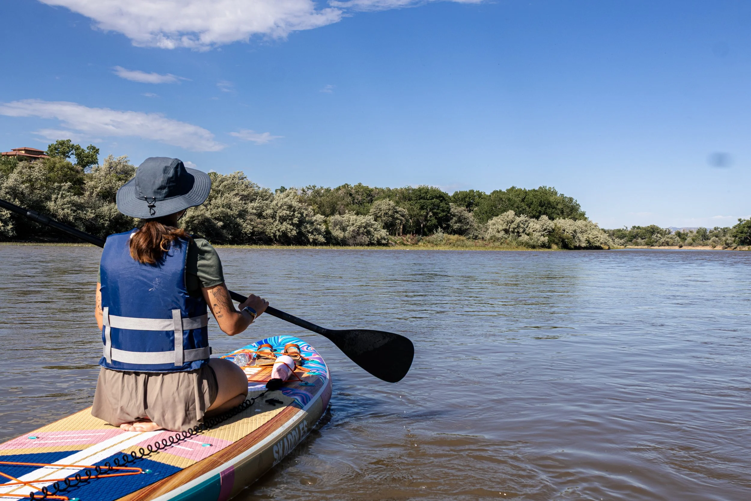 A person kneeling on a paddleboard on a calm river, wearing a wide-brimmed hat, a life vest, and beige shorts, holding a paddle, with dense trees and blue sky in the background.