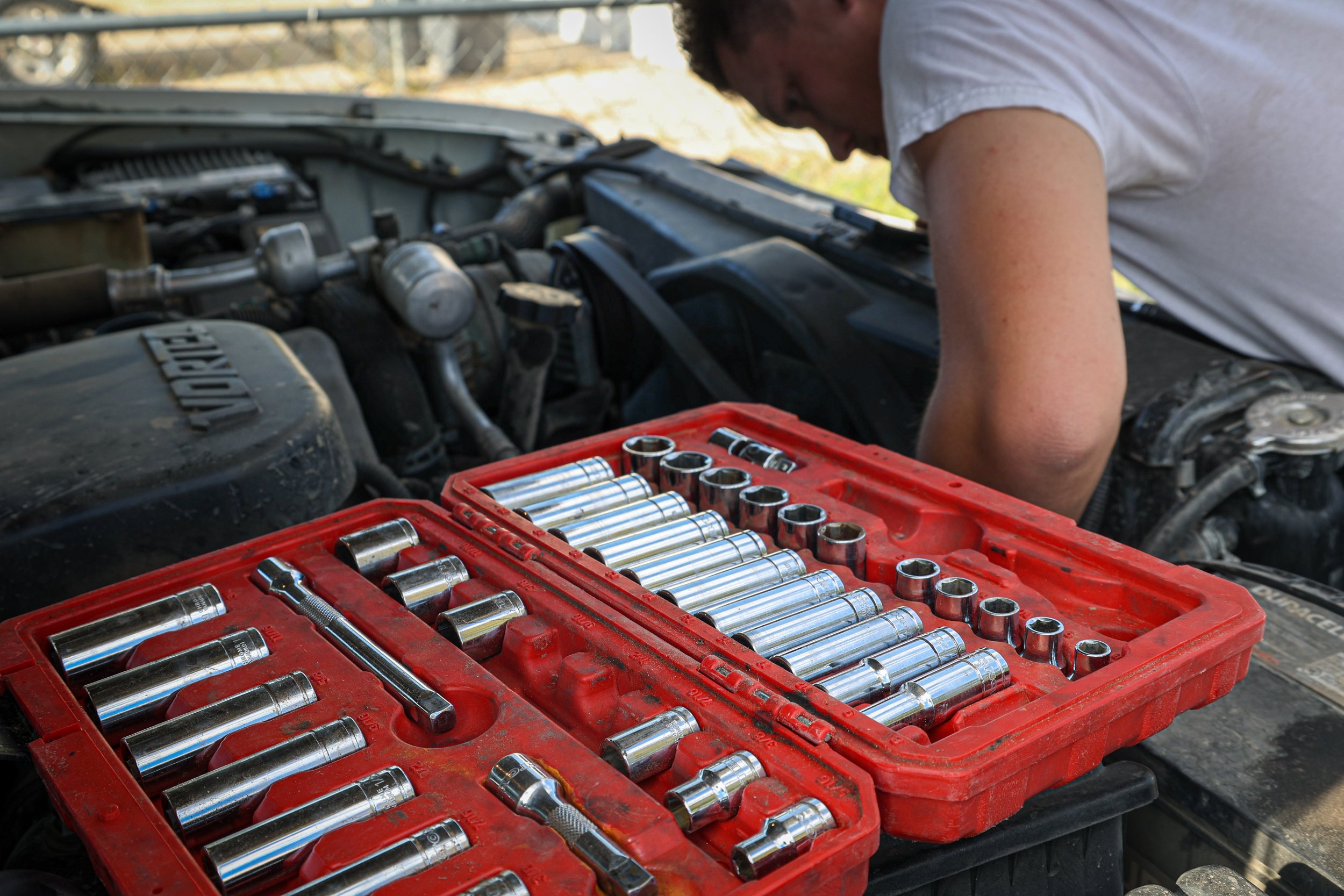 A person working on a car engine with a red toolbox containing socket wrench tools in the foreground.