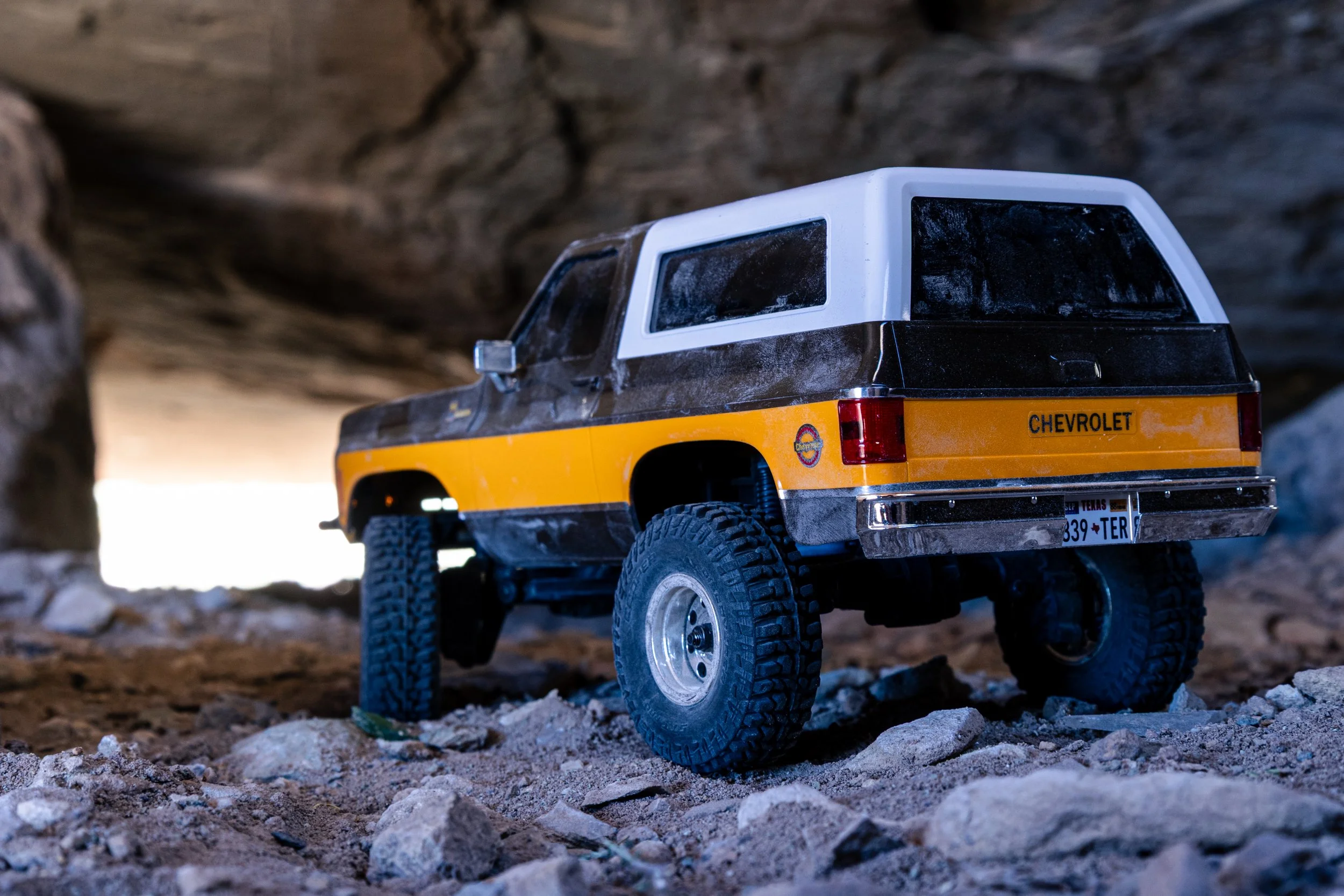 A toy off-road truck with a black and white roof, yellow body, and large tires, positioned on rocky terrain inside a cave or tunnel.