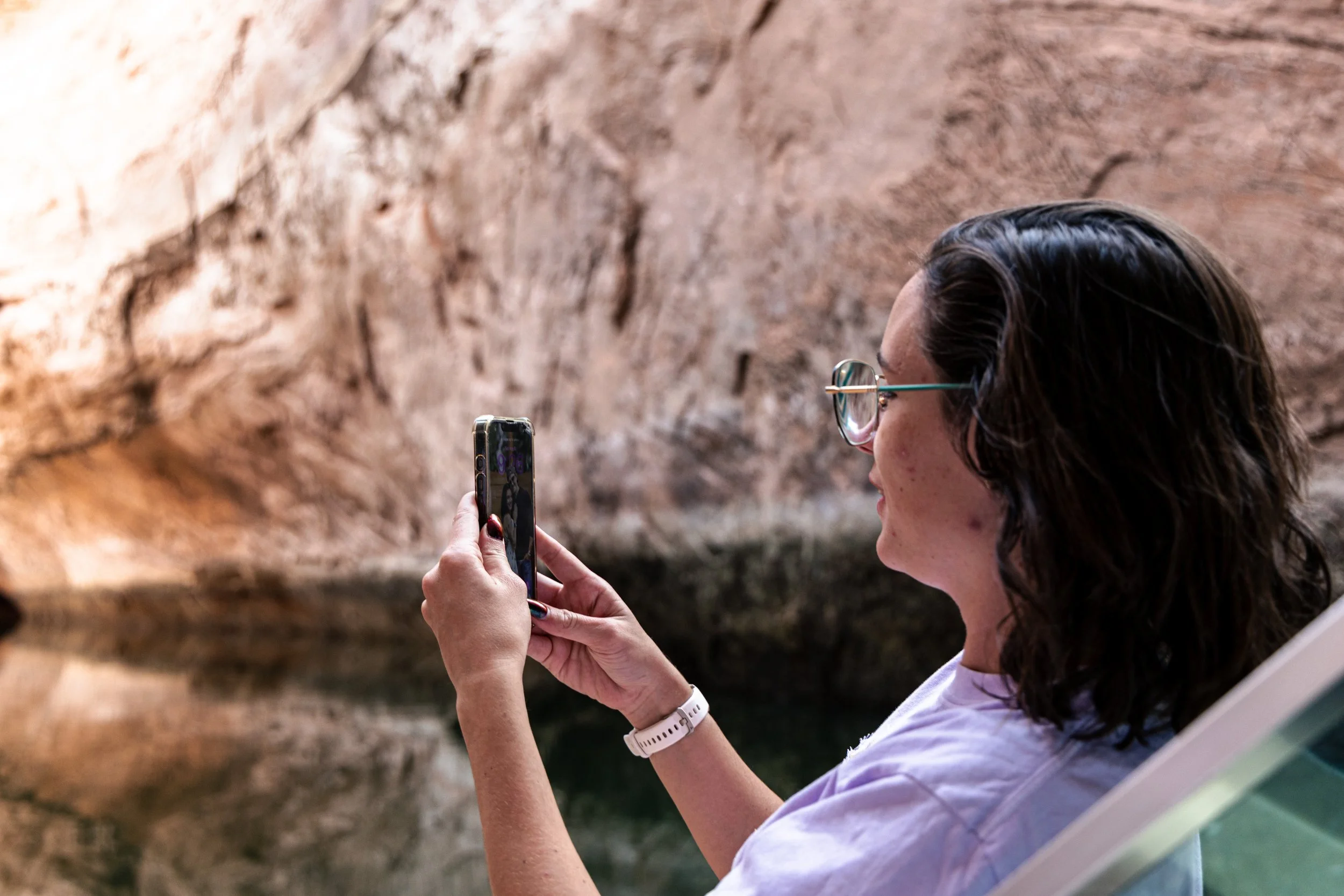 A woman with glasses and dark hair taking a photo on her phone inside a cave or tunnel with rocky walls.