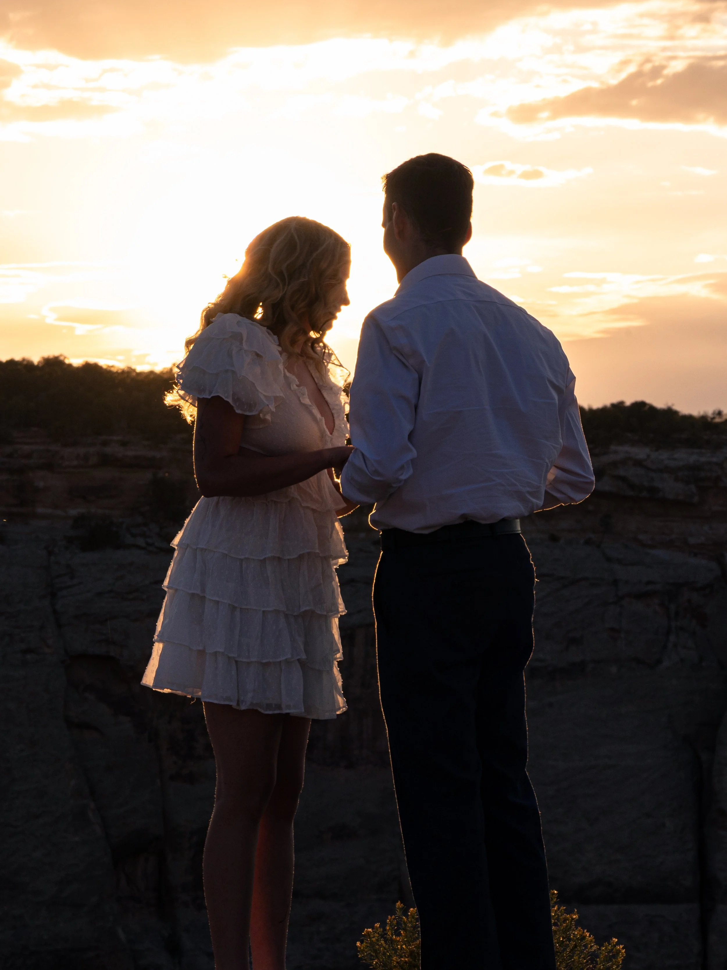A couple standing close together outdoors during sunset, facing each other with the sky illuminated behind them.