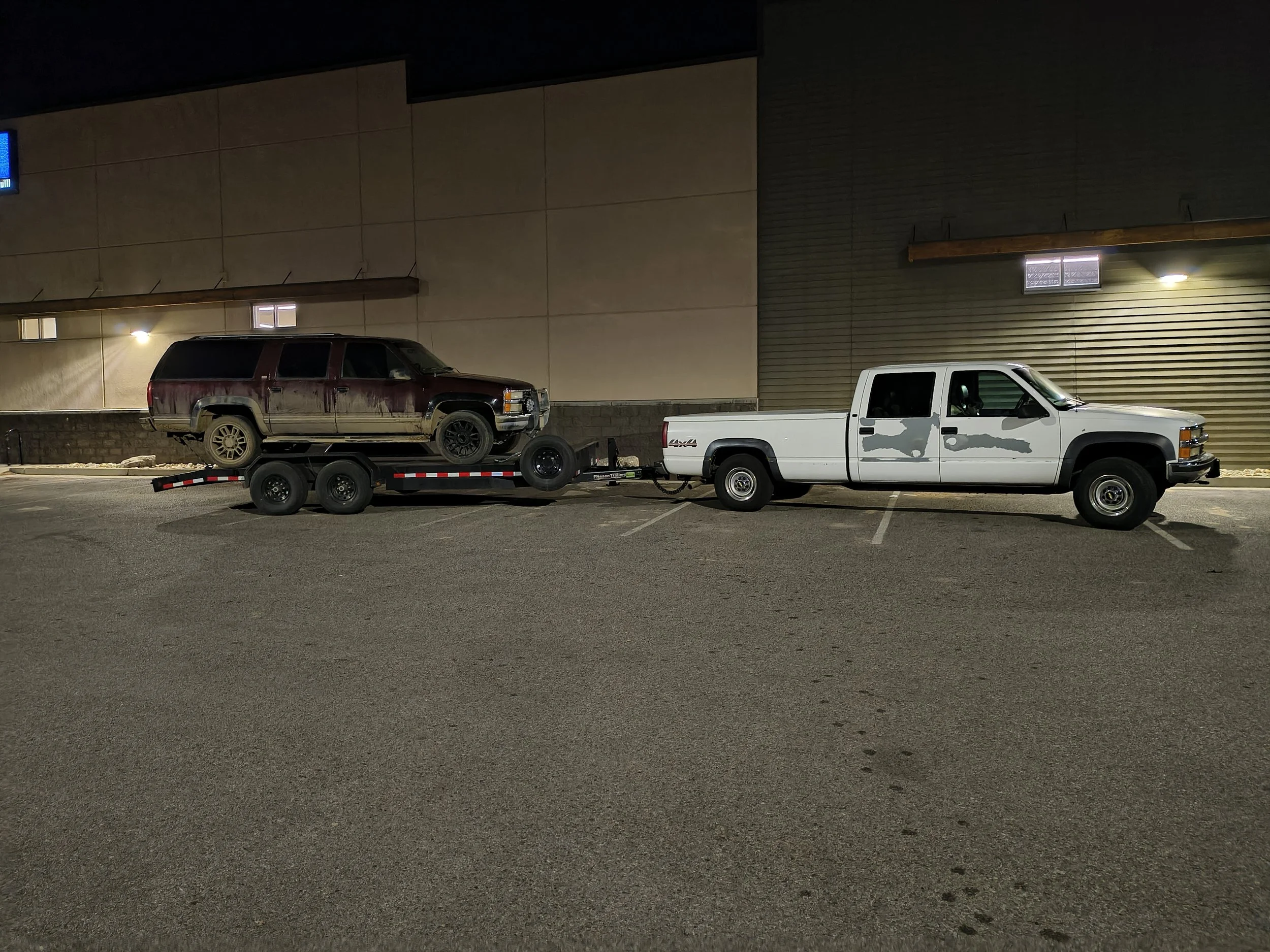 Nighttime scene of a parking lot with a white pickup truck towing a trailer carrying a rusty red SUV, with a beige building with small windows and exterior lights in the background.