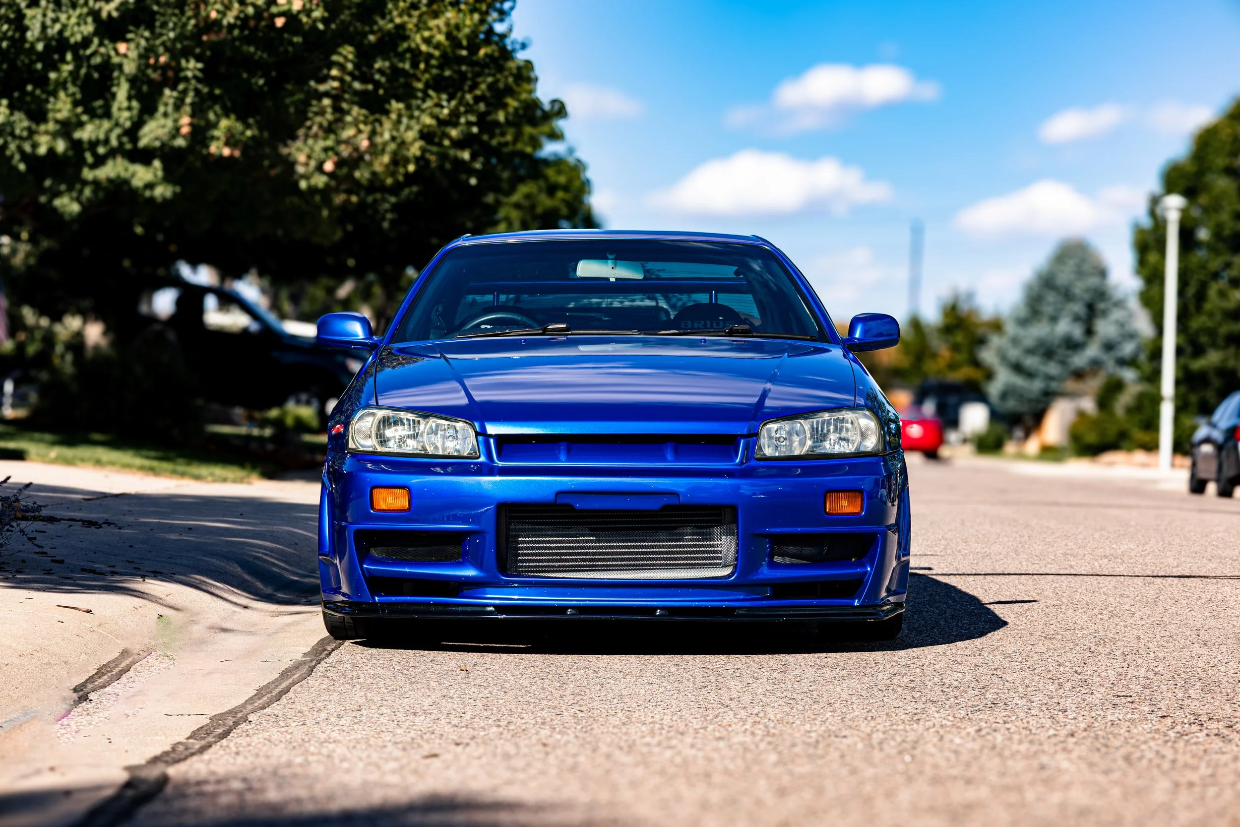 Front view of a blue sports car parked on a street with trees and a blue sky in the background.