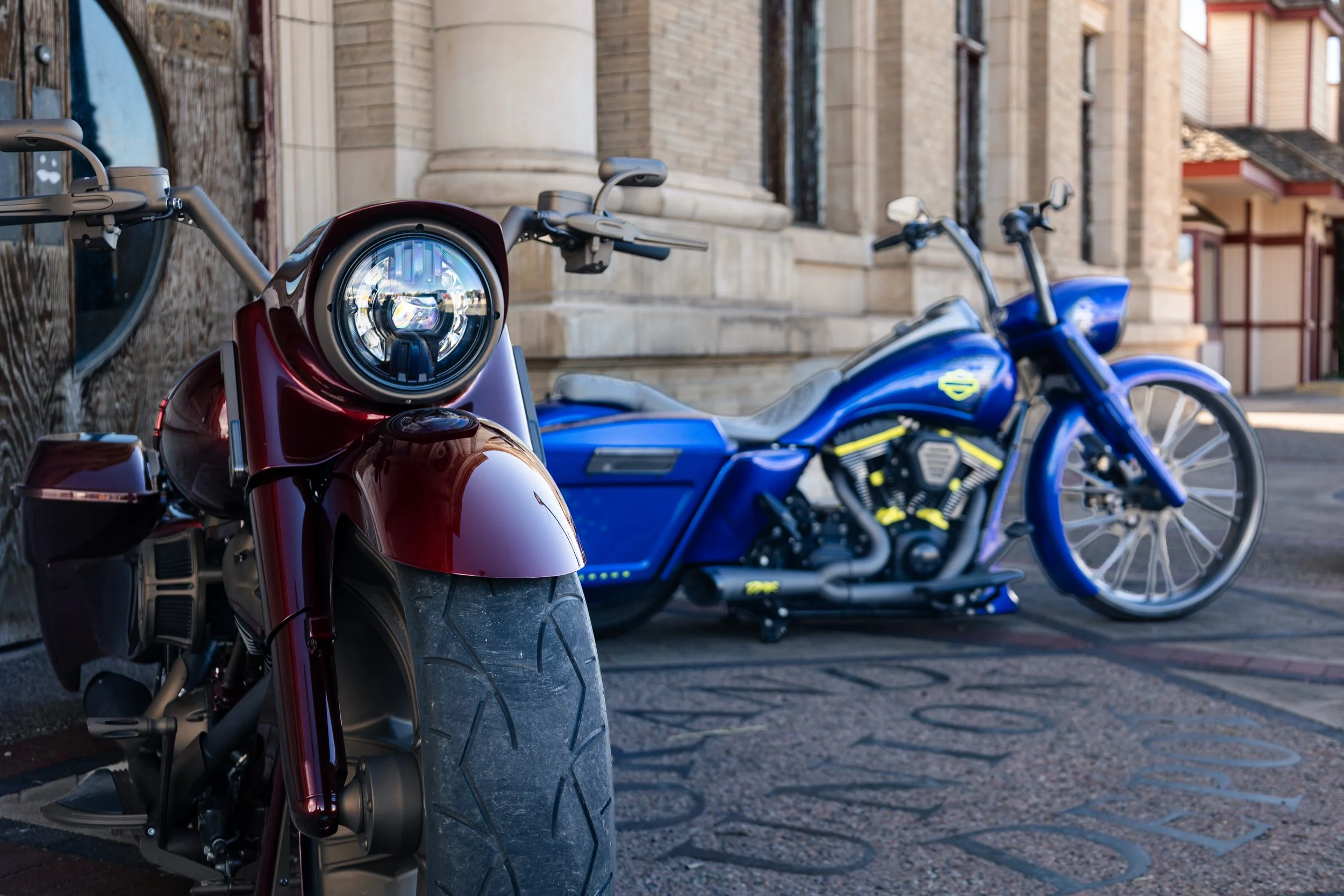 A close-up of a red motorcycle with a round headlight, parked on a city street near a brick building. In the background, there are two blue motorcycles, also parked along the street.