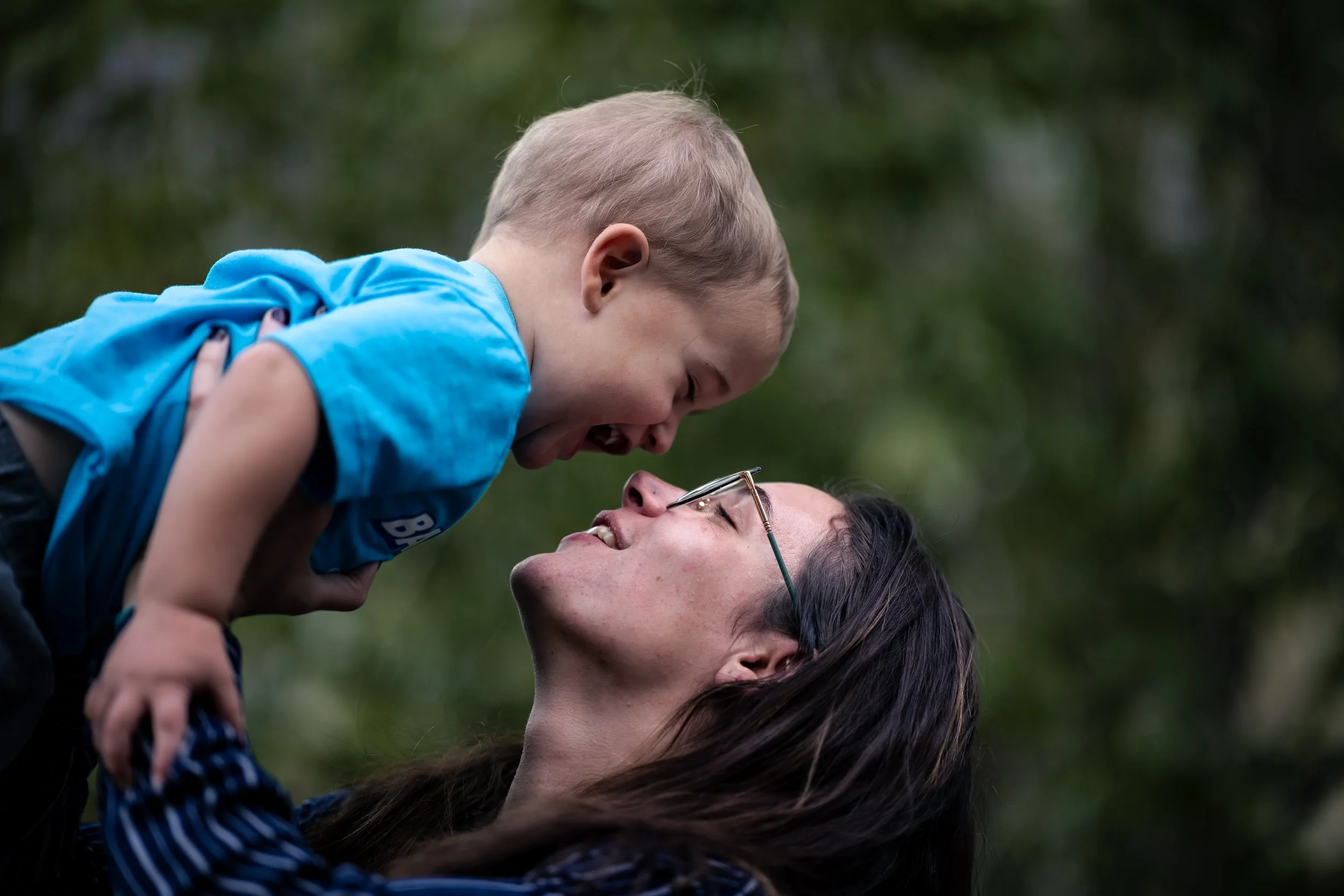 A woman lying on her back outdoors with her eyes closed, smiling while a young boy with glasses and light hair leans down from above and kisses her on the lips.