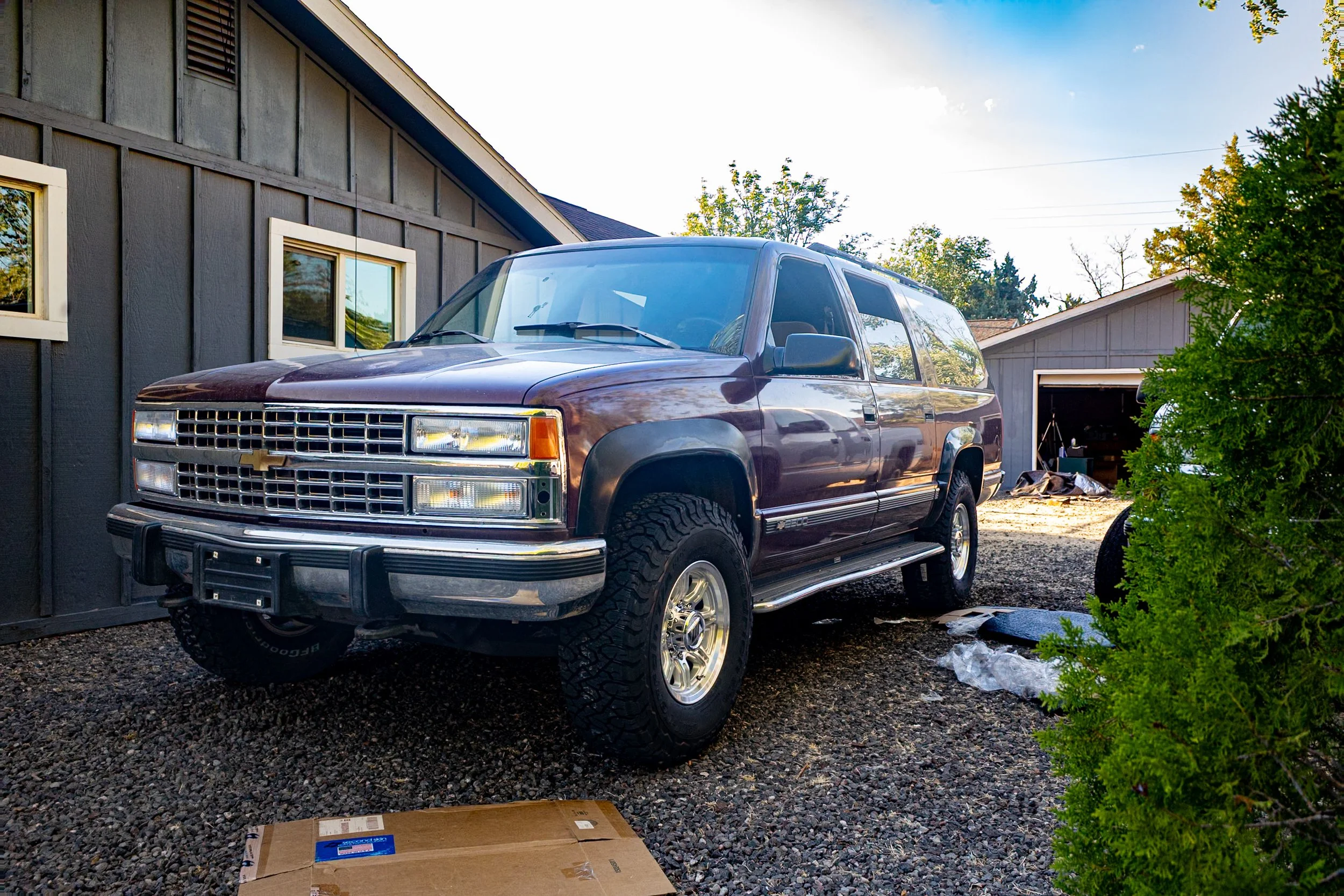 A maroon Chevrolet SUV parked on a gravel driveway in front of a gray house with white-framed windows and a garage. There is a cardboard box on the ground in the foreground and some trash in the garage. Trees and a clear sky are in the background.