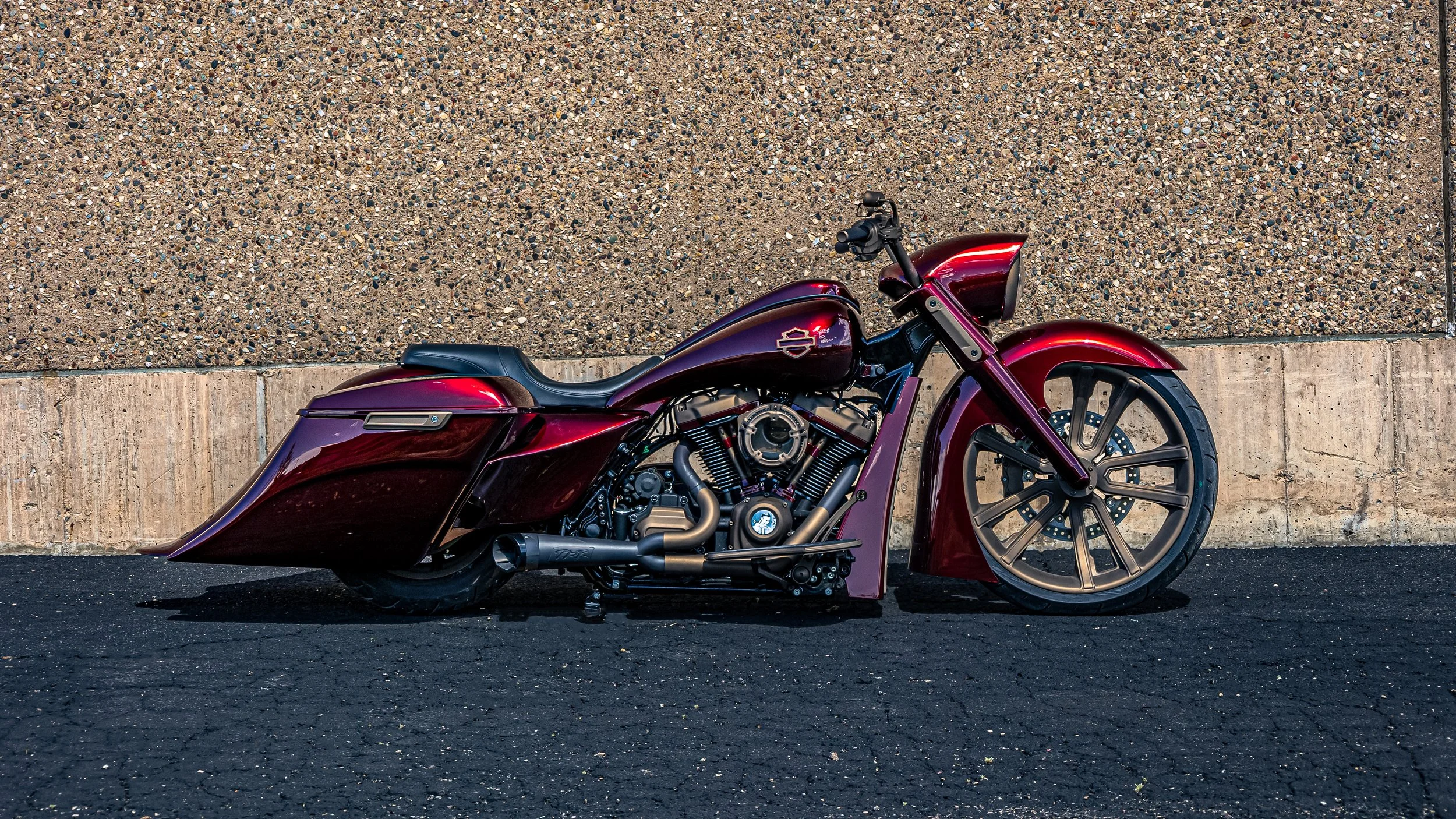 A custom maroon motorcycle with black accents parked on black asphalt against a textured concrete wall.