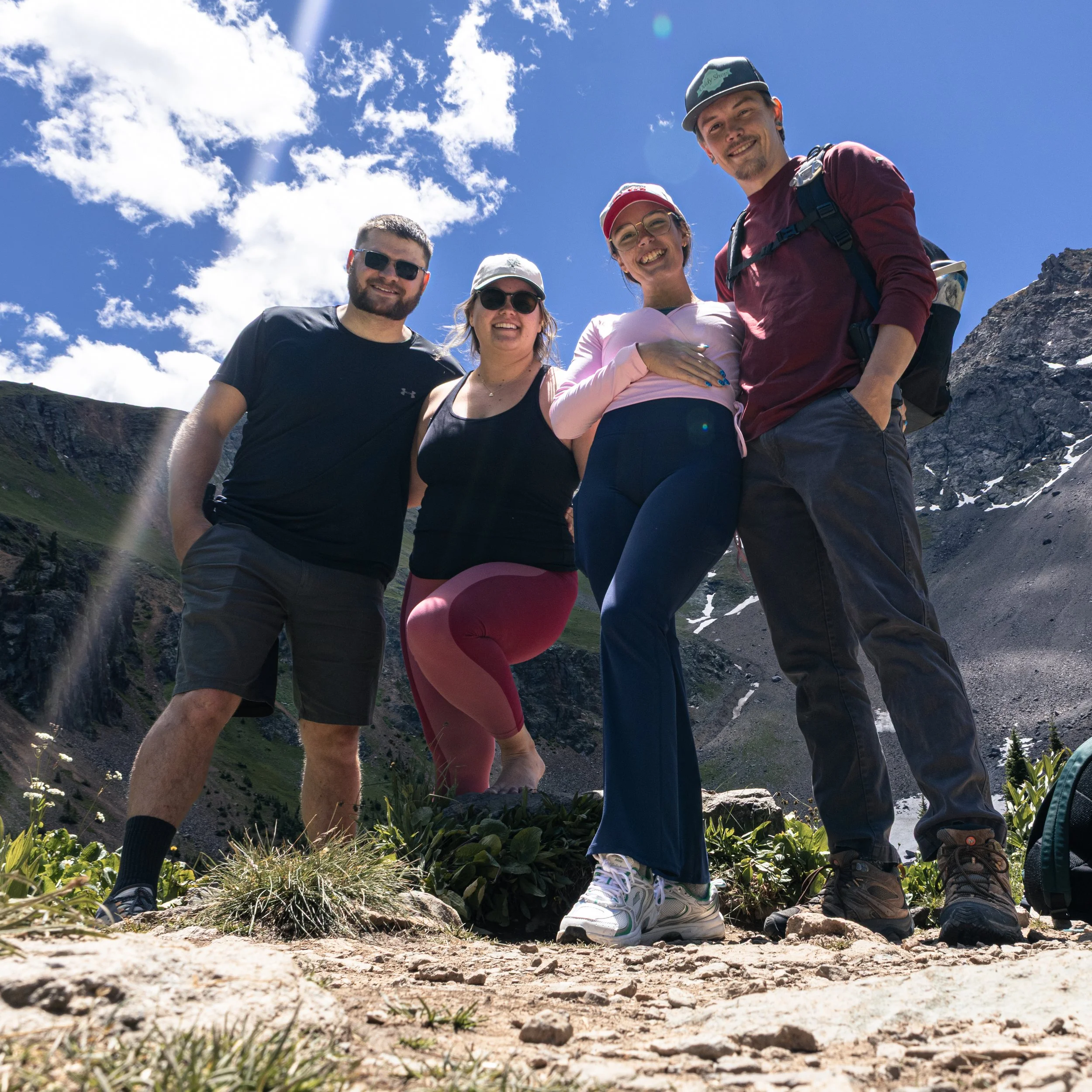 A group of five friends standing on a mountain trail under blue sky with clouds, smiling and wearing casual outdoor clothing, with mountains and rocky terrain in the background.