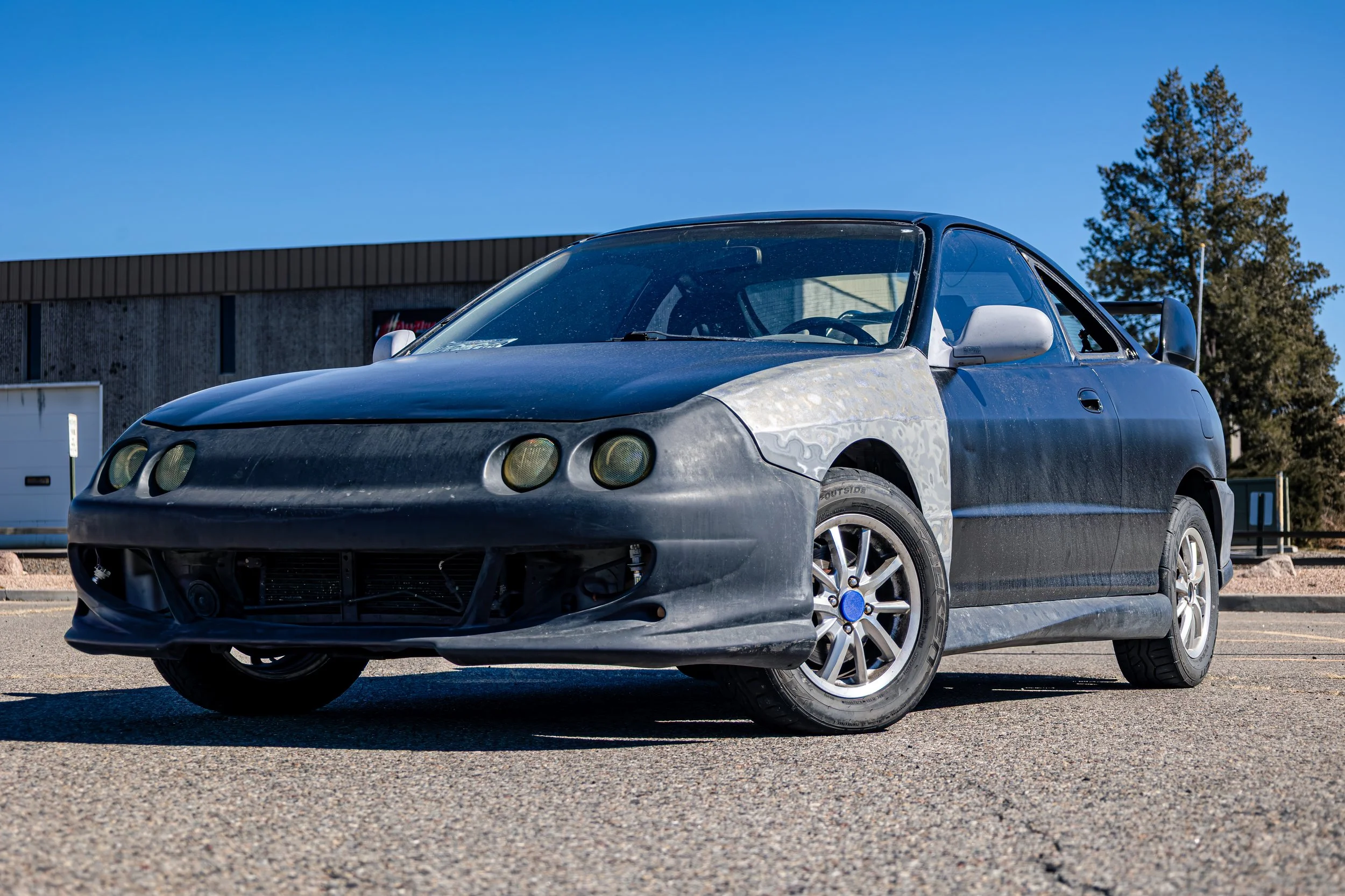 A partially modified sports car with a matte black front bumper and hood, silver side mirror, and custom wheels parked outside near a building and trees under a blue sky.
