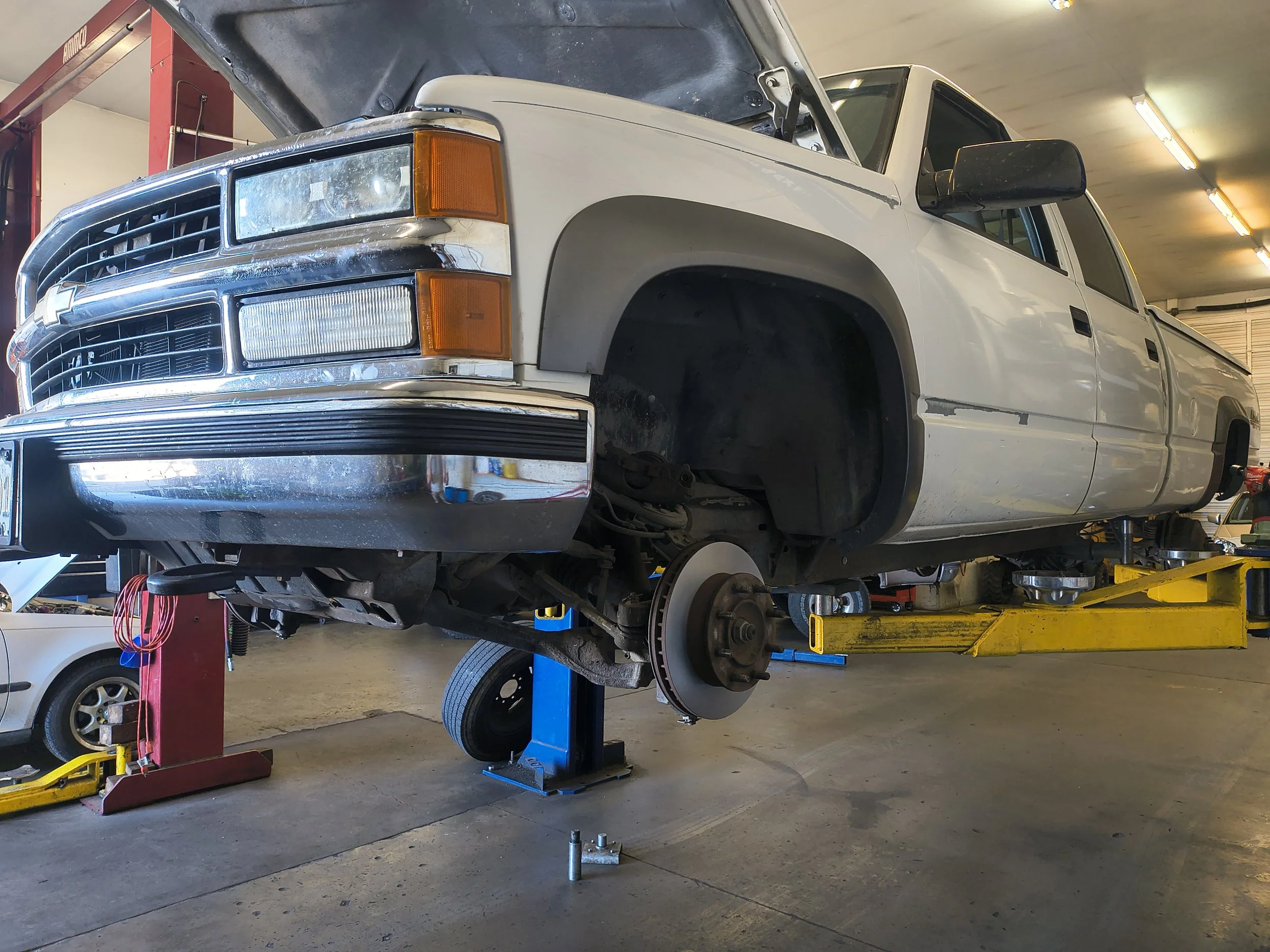White pickup truck on a hydraulic lift at an auto repair shop, with the front tire removed, exposing the brake and suspension components.