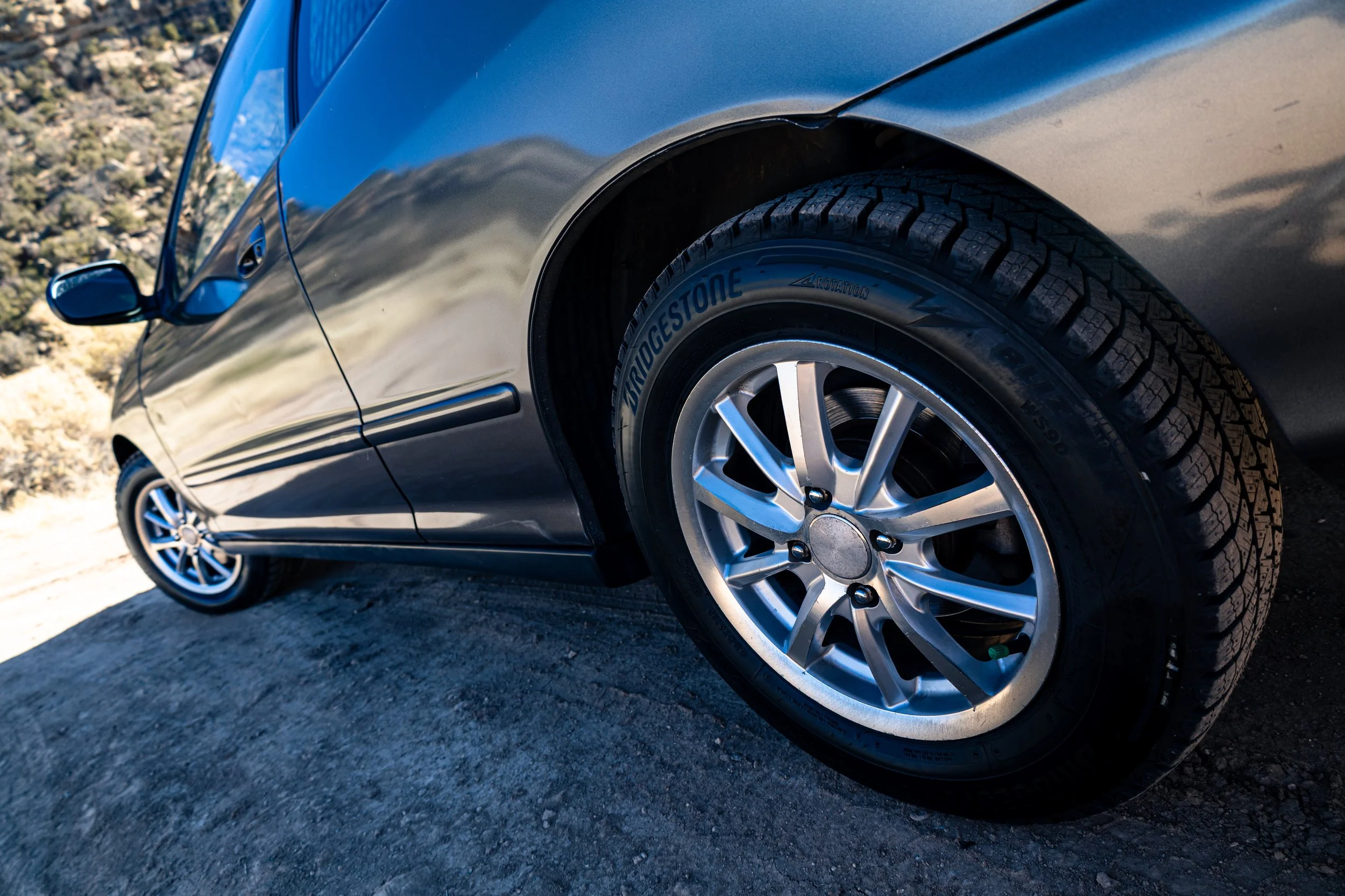 Close-up of a silver car's front wheel and tire on a gravel or asphalt surface, with part of the car's side and door visible.
