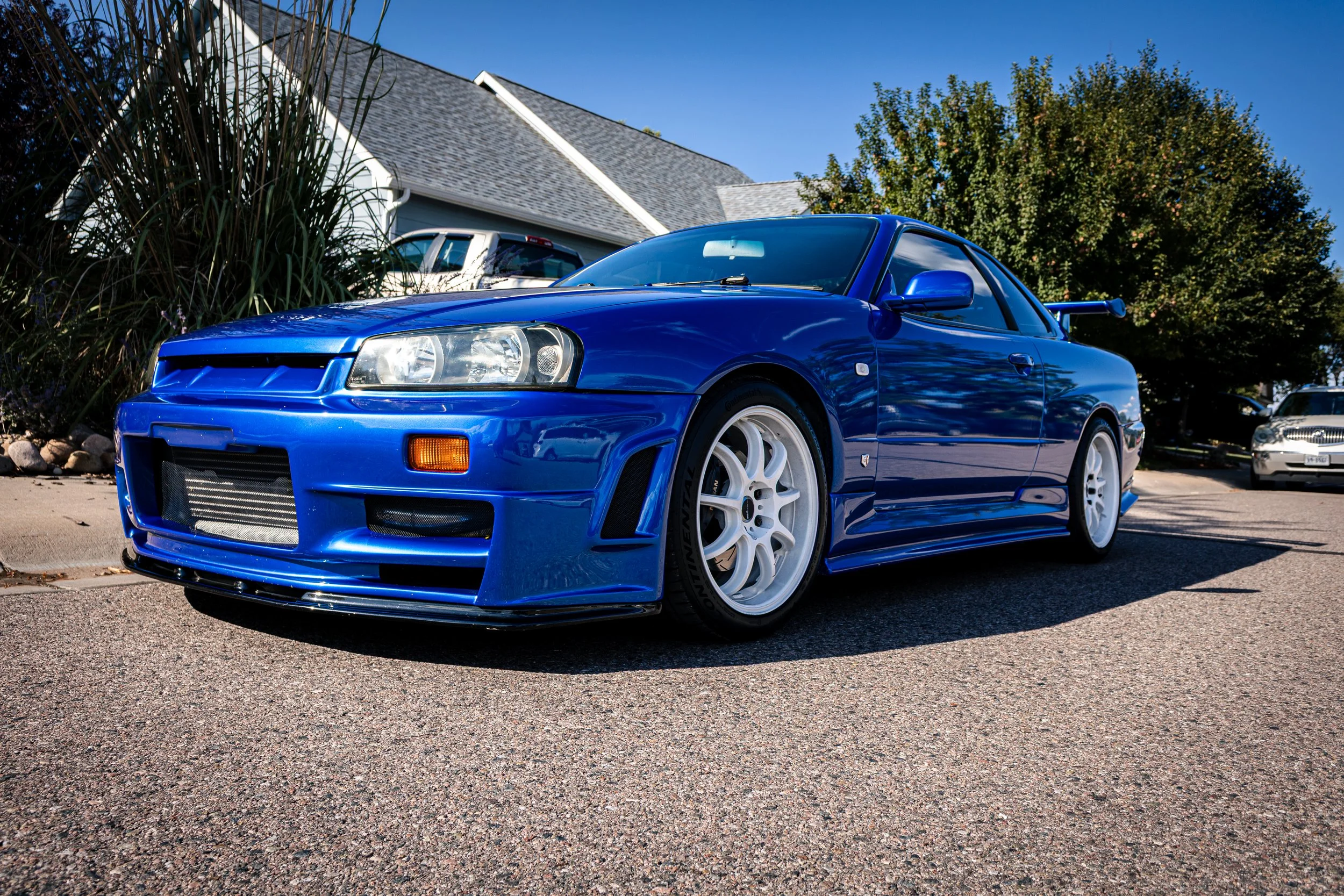 A bright blue sports car with white wheels parked in a driveway, with a blue house and trees in the background under a clear blue sky.