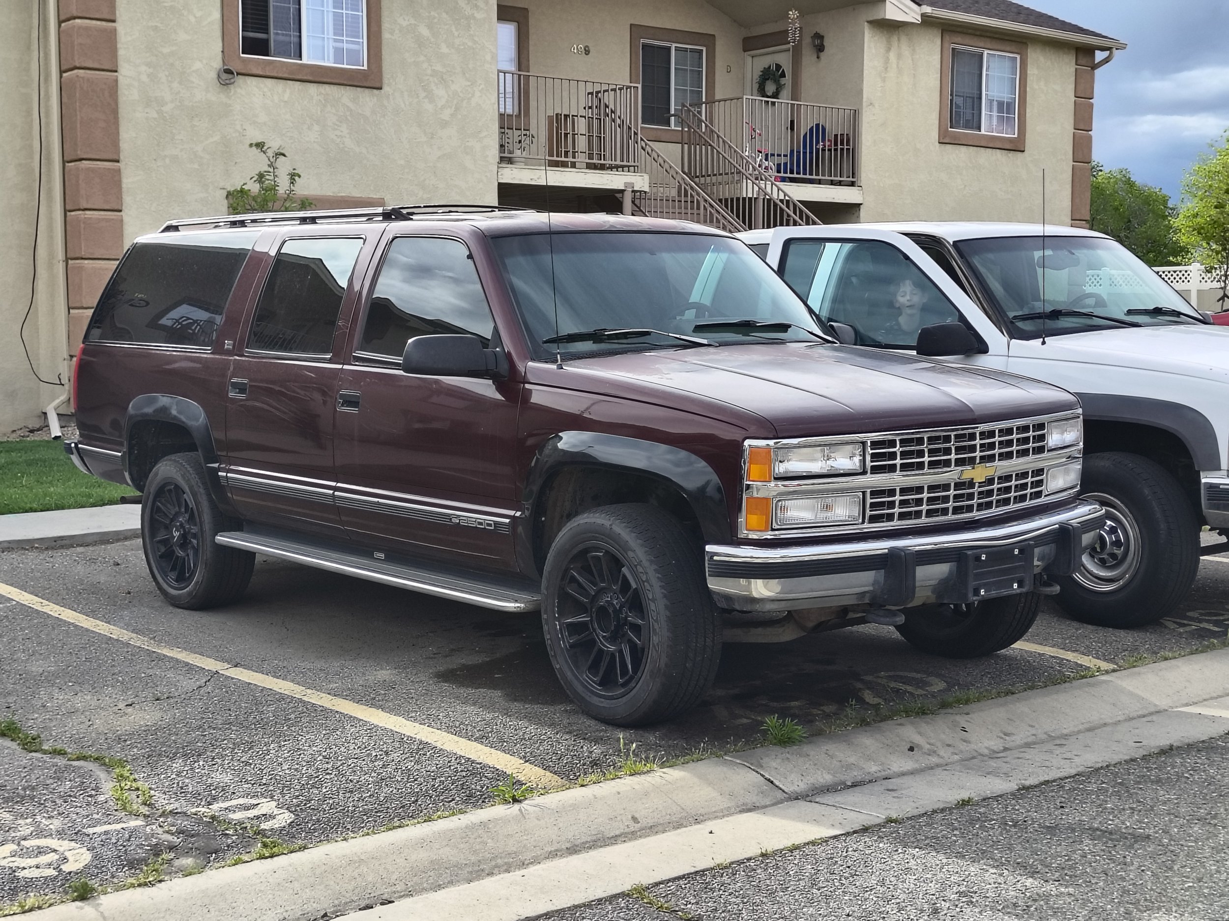 Two parked cars, a maroon SUV and a white van, in front of an apartment building with stairs, windows, and balconies, during daytime.