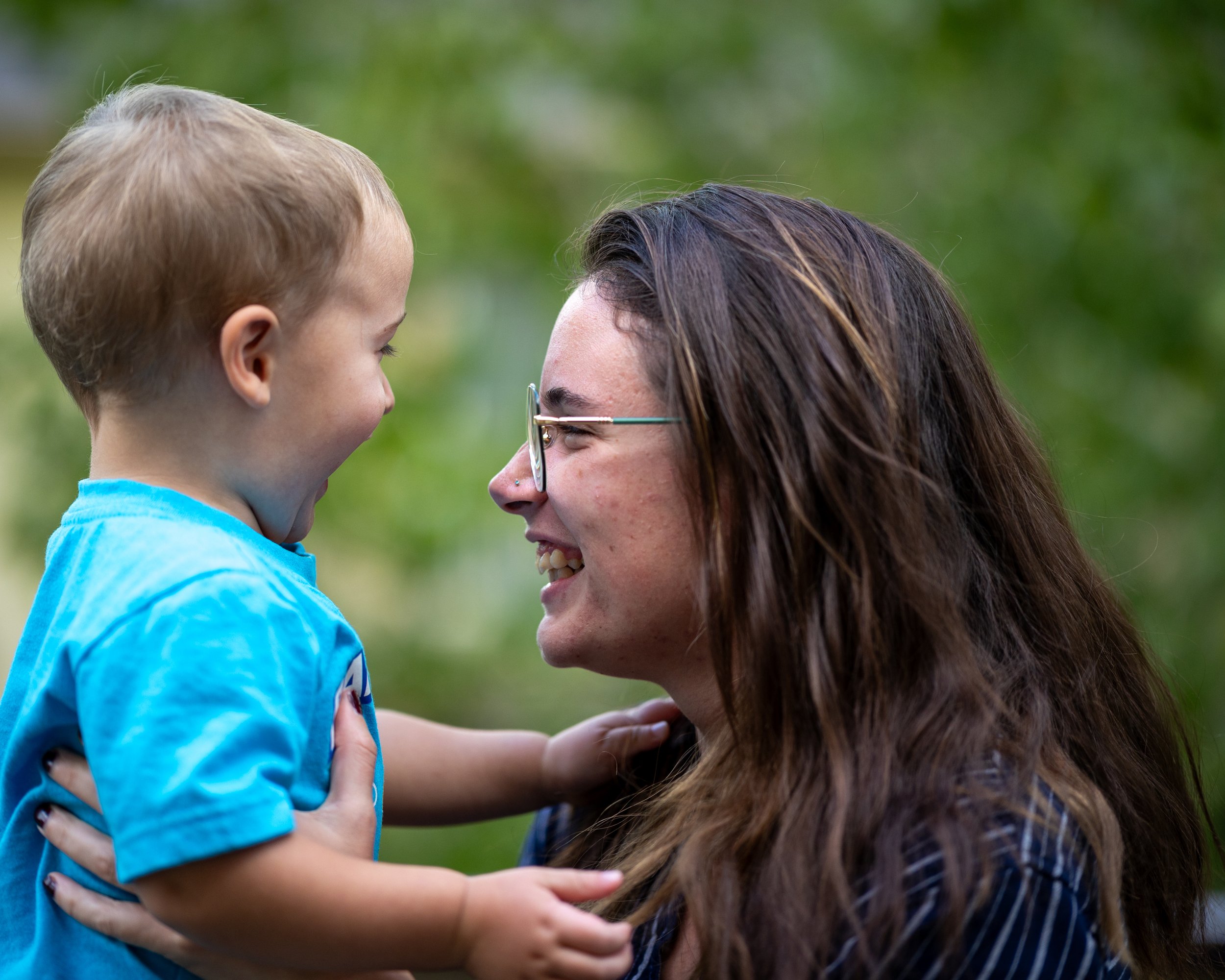 A woman with long brown hair and glasses smiling while hugging and leaning her forehead against a young boy with short blonde hair, wearing a bright blue shirt, in an outdoor green setting.