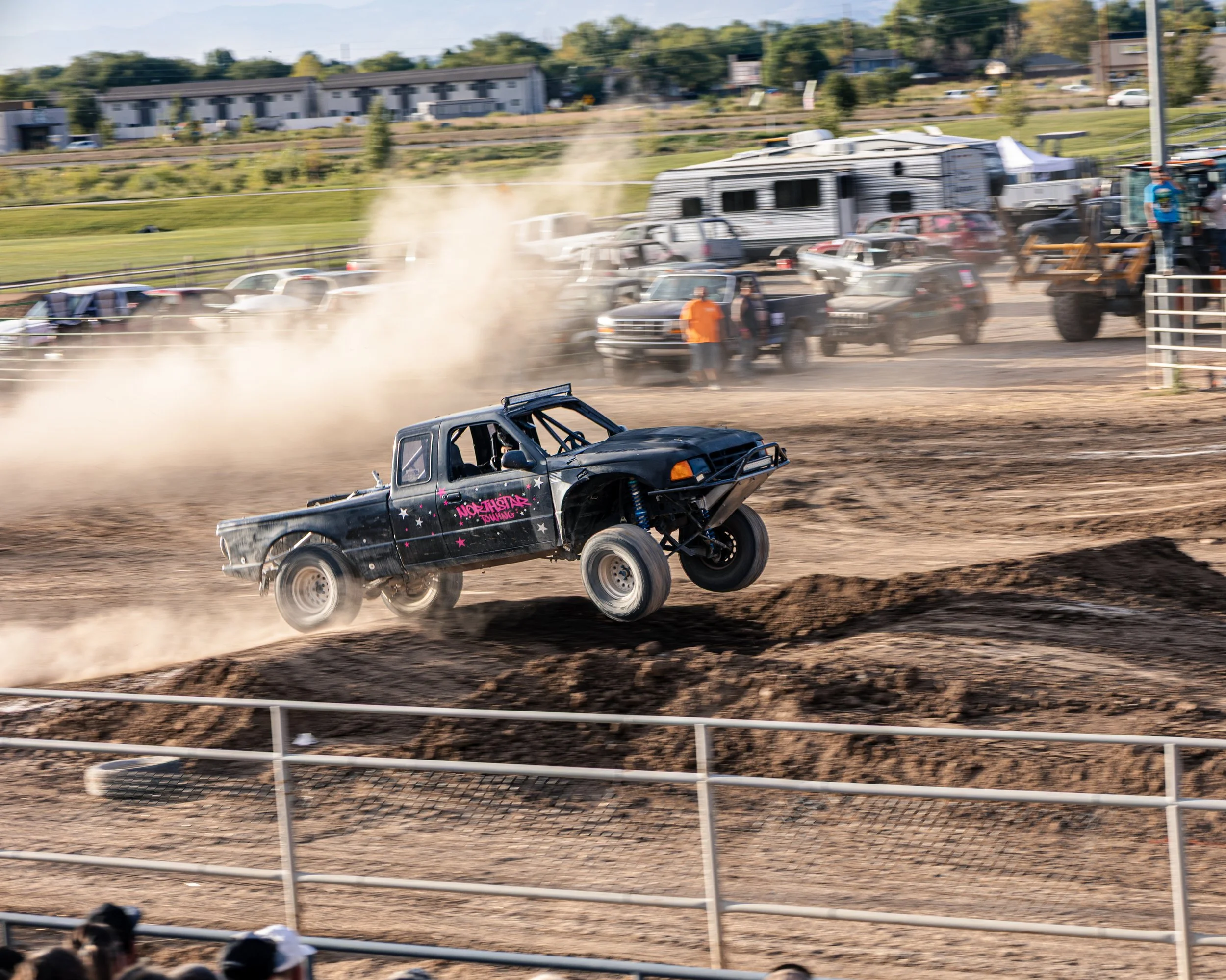 A dirt track race with a black off-road vehicle in mid-air during a jump. The vehicle has purple and pink graphics and the words 'North Shasta Racing'. There are spectators watching behind a safety fence, and in the background, cars, trailers, and tr