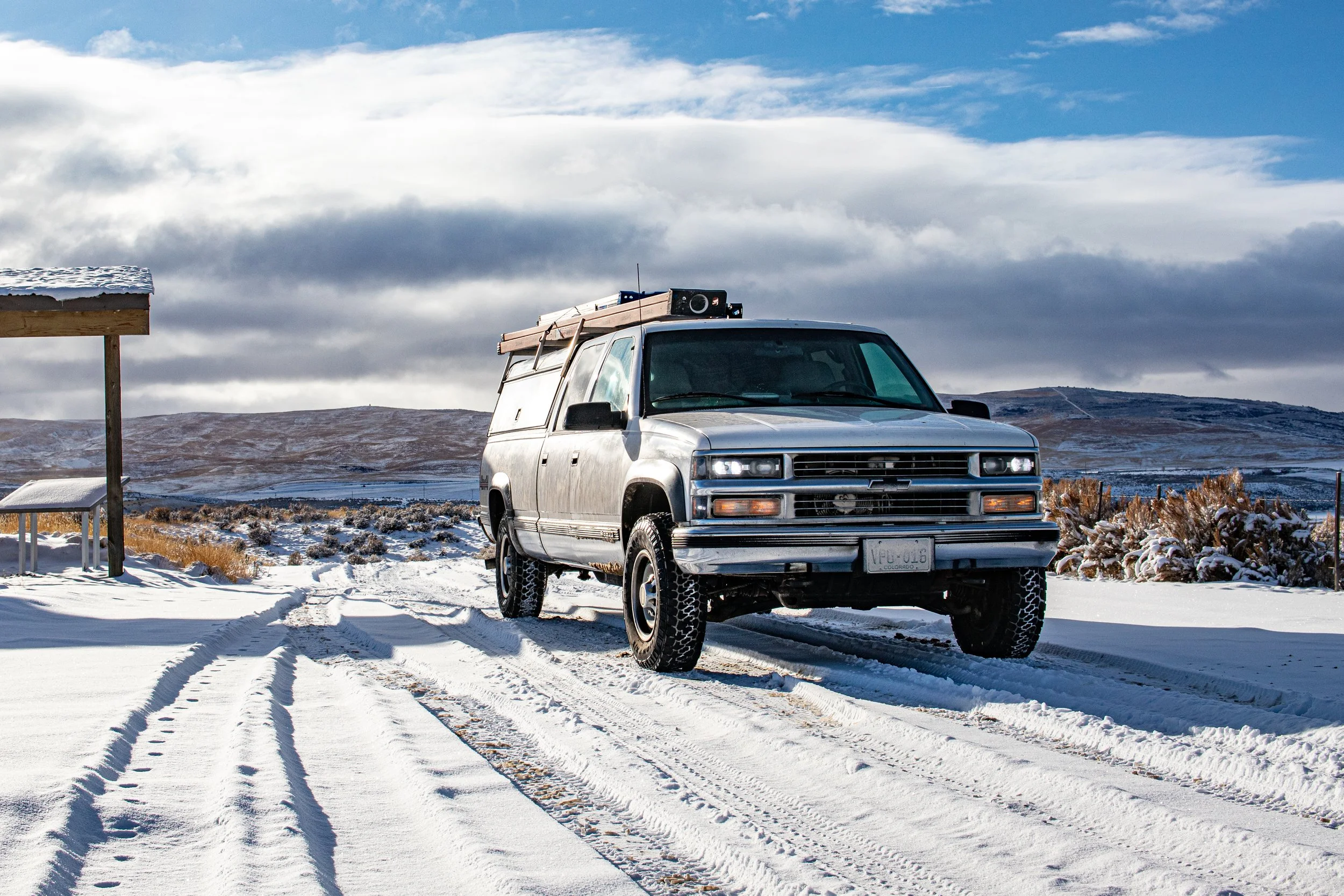 A silver SUV parked on snow-covered ground with tire tracks, under a cloudy sky, in a rural area with distant hills and a small structure.