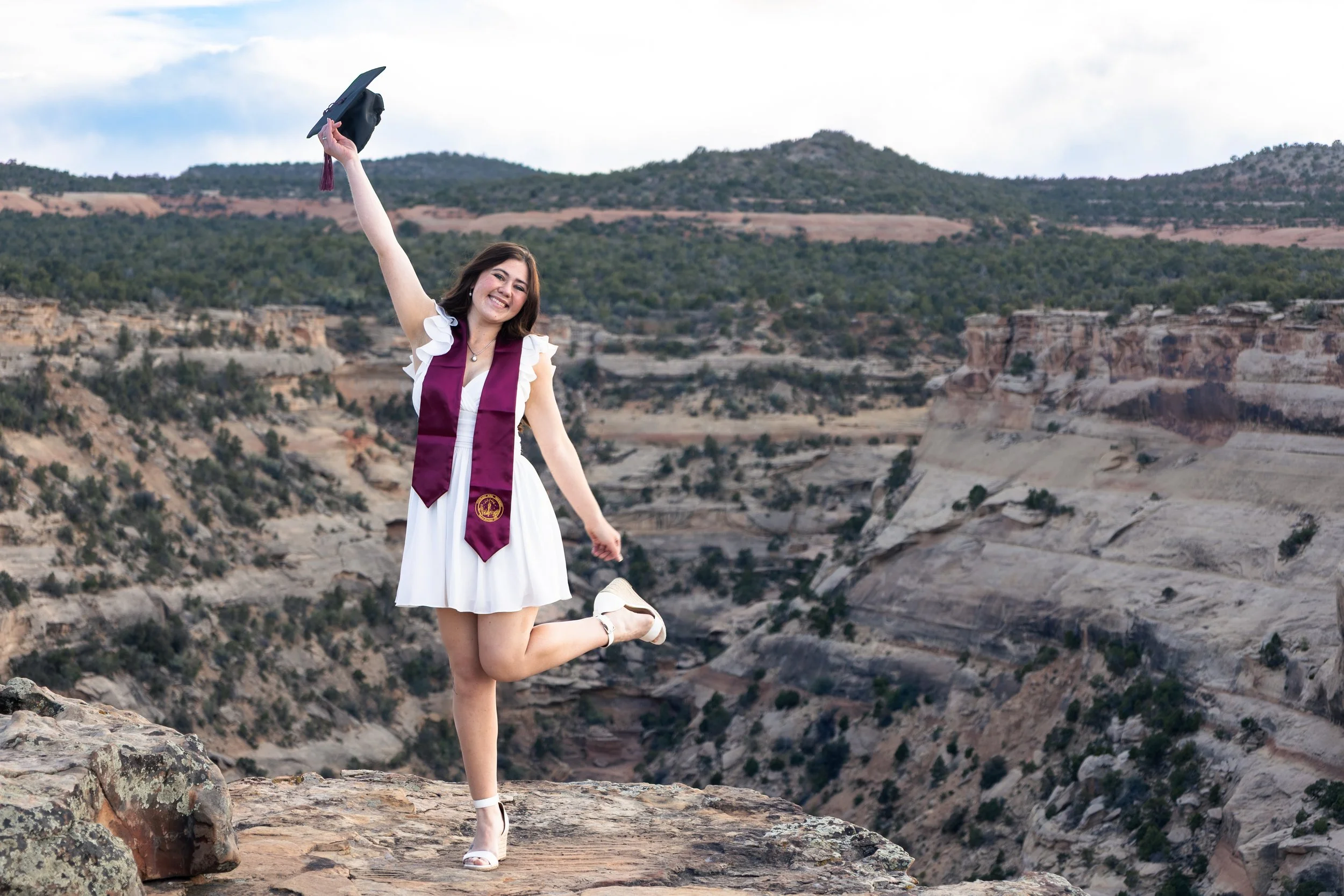 A young woman in a white dress and white sandals celebrating her graduation, holding her cap in the air, standing on a rocky ledge with a canyon landscape and hills in the background.