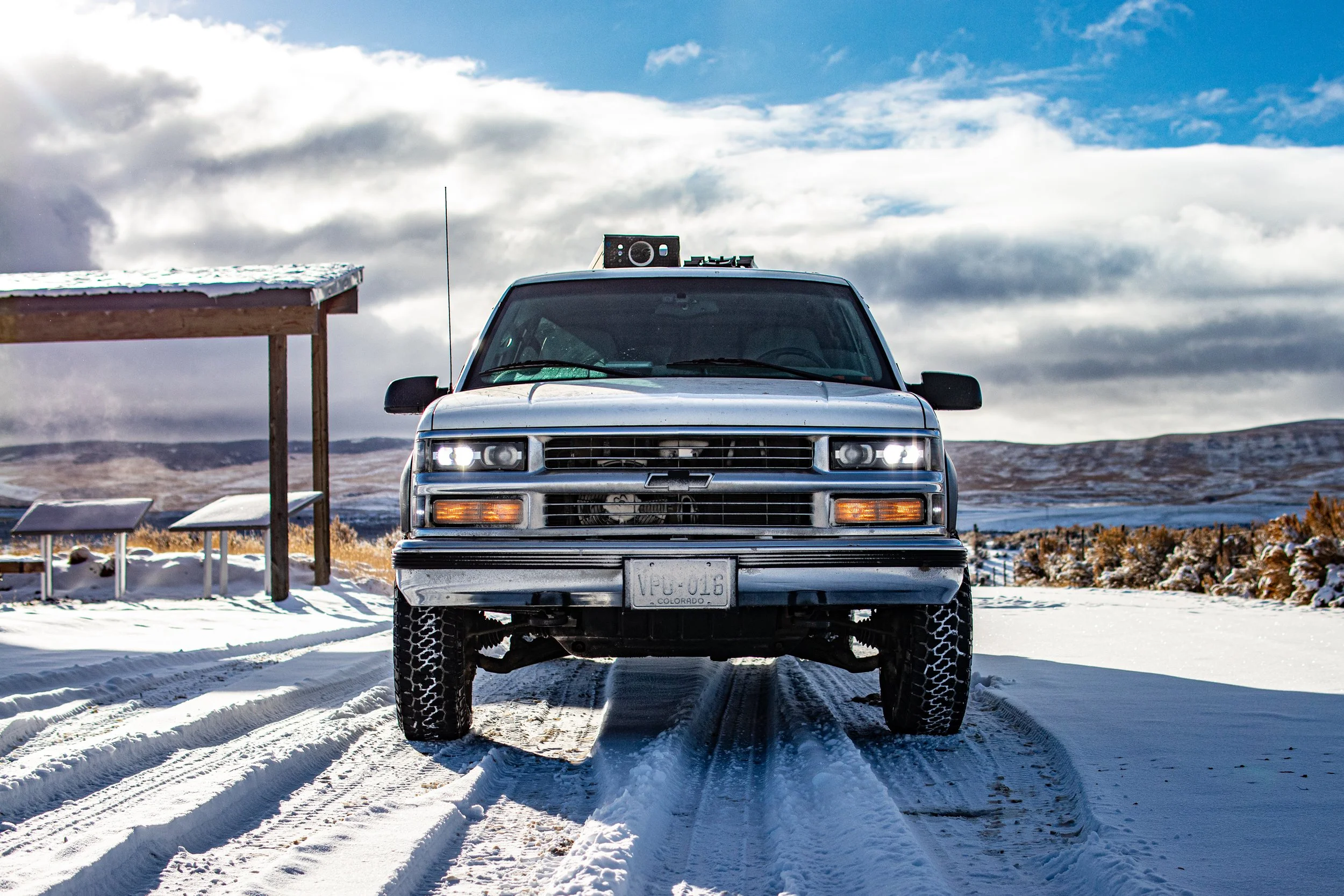 A silver Chevrolet SUV parked on snow-covered ground with tire tracks, in front of a scenic landscape with cloudy sky, mountains, and a snow-covered structure on the left.
