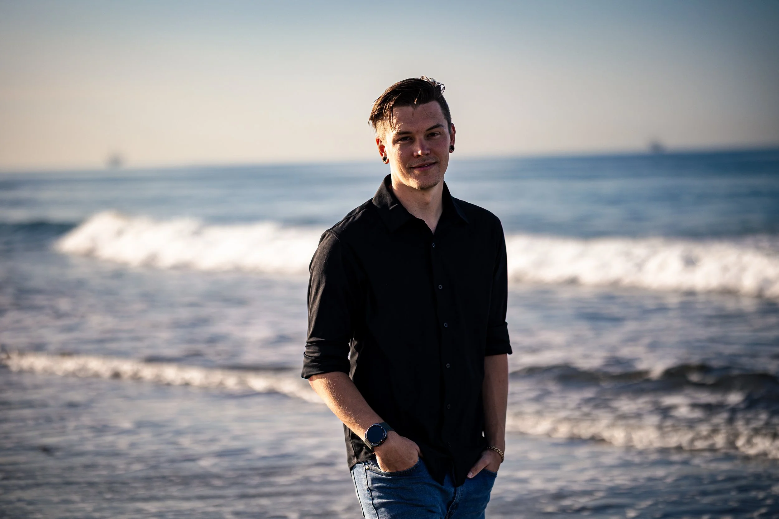 A young man in a black shirt and jeans standing on the beach near the ocean with waves in the background, during late afternoon or early evening.