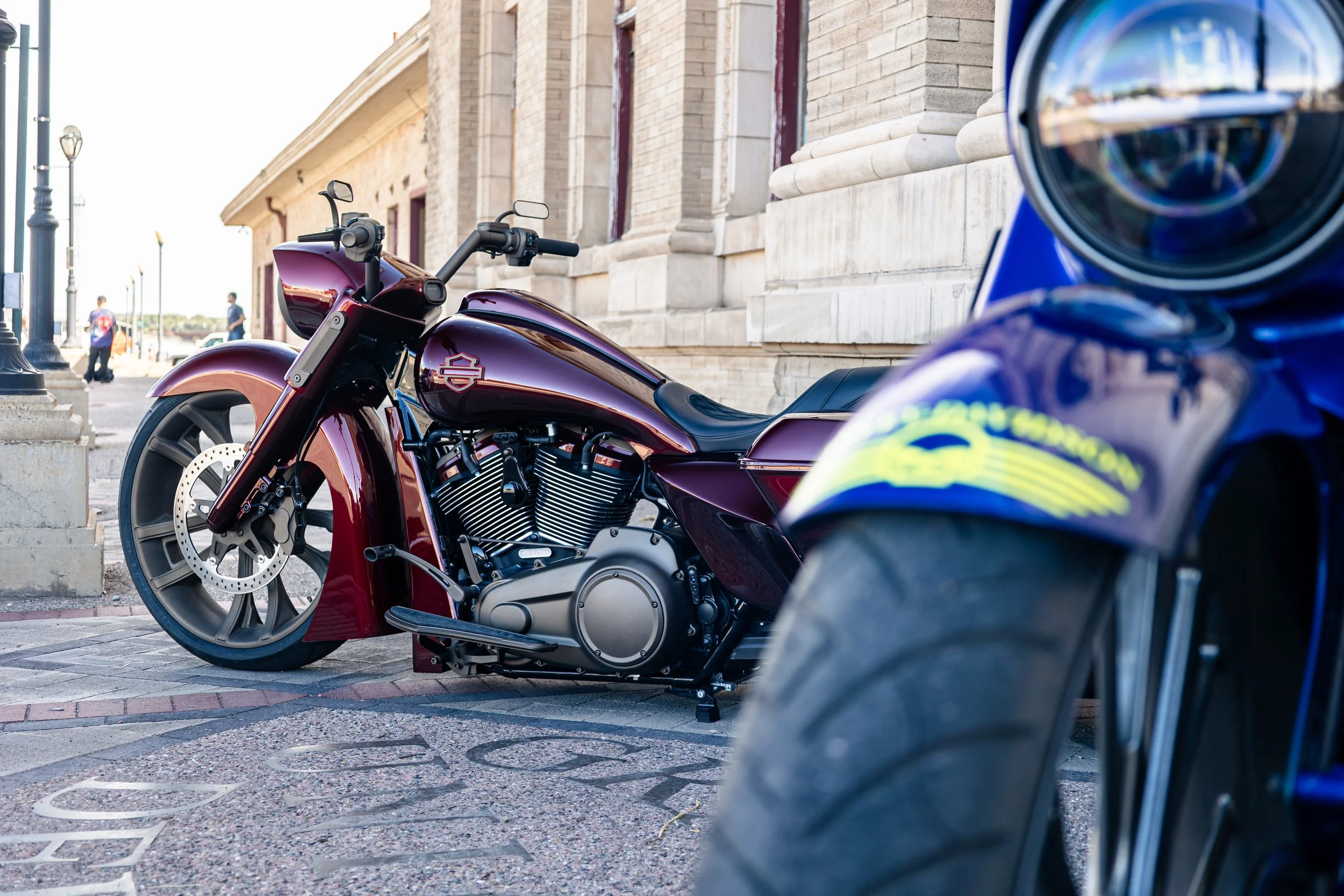 Close-up of a dark red motorcycle parked on a city street with a historic building in the background and a partially visible blue motorcycle in the foreground.