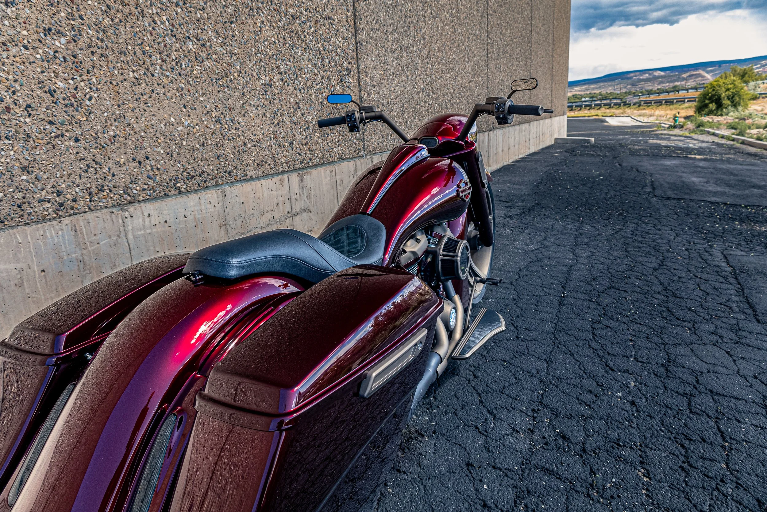 A red and black motorcycle parked next to a textured concrete wall under a cloudy sky with distant mountains and trees in the background.