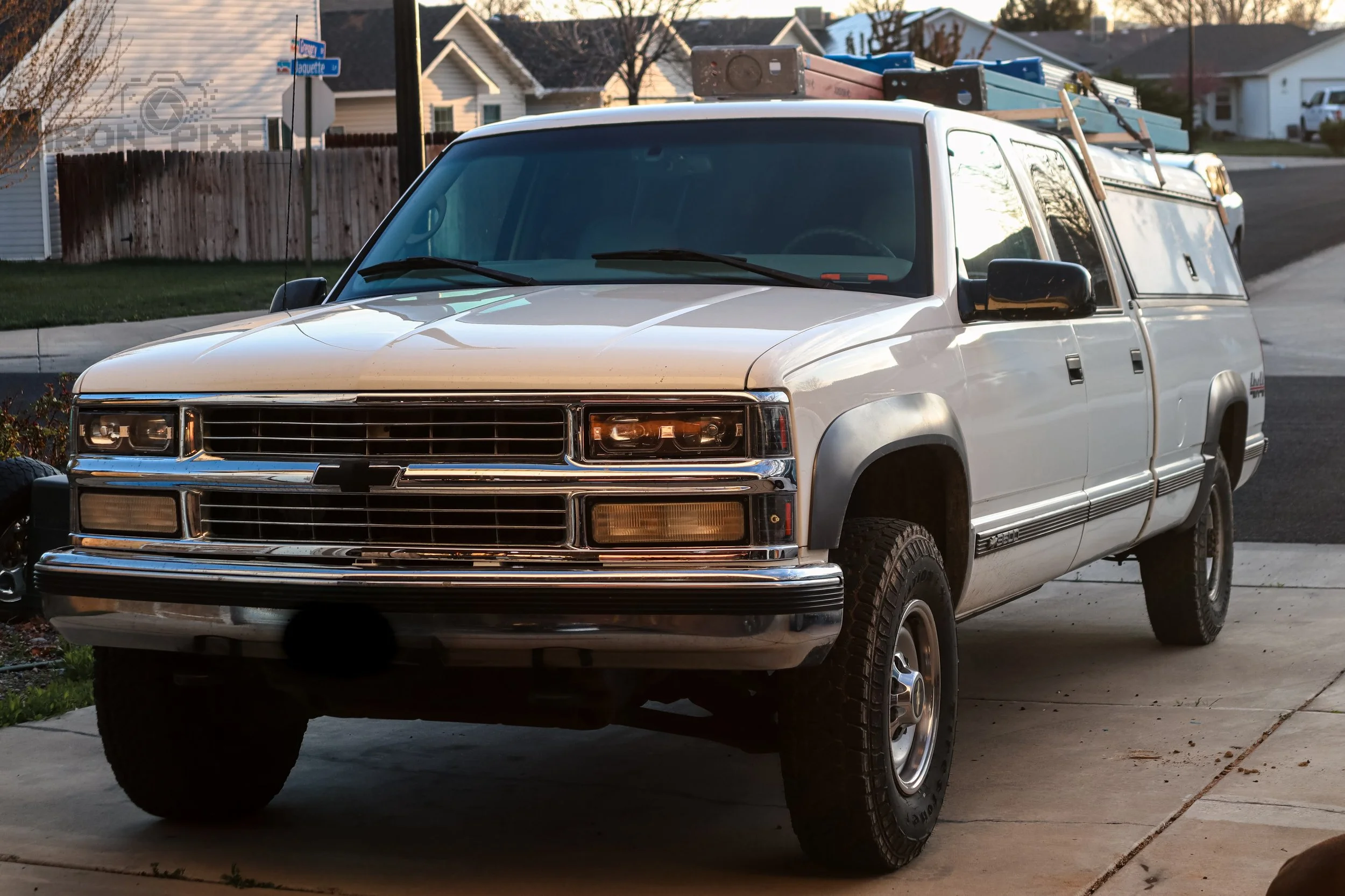 A white Chevrolet pickup truck with a camper shell parked on a driveway in a suburban neighborhood. Ladders are mounted on the roof of the truck.