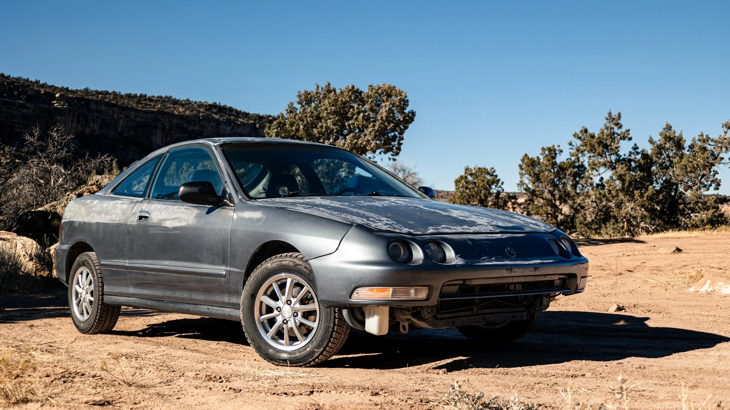 A weathered silver hatchback car parked on a dirt surface in a desert landscape with trees and hills in the background under a clear blue sky.