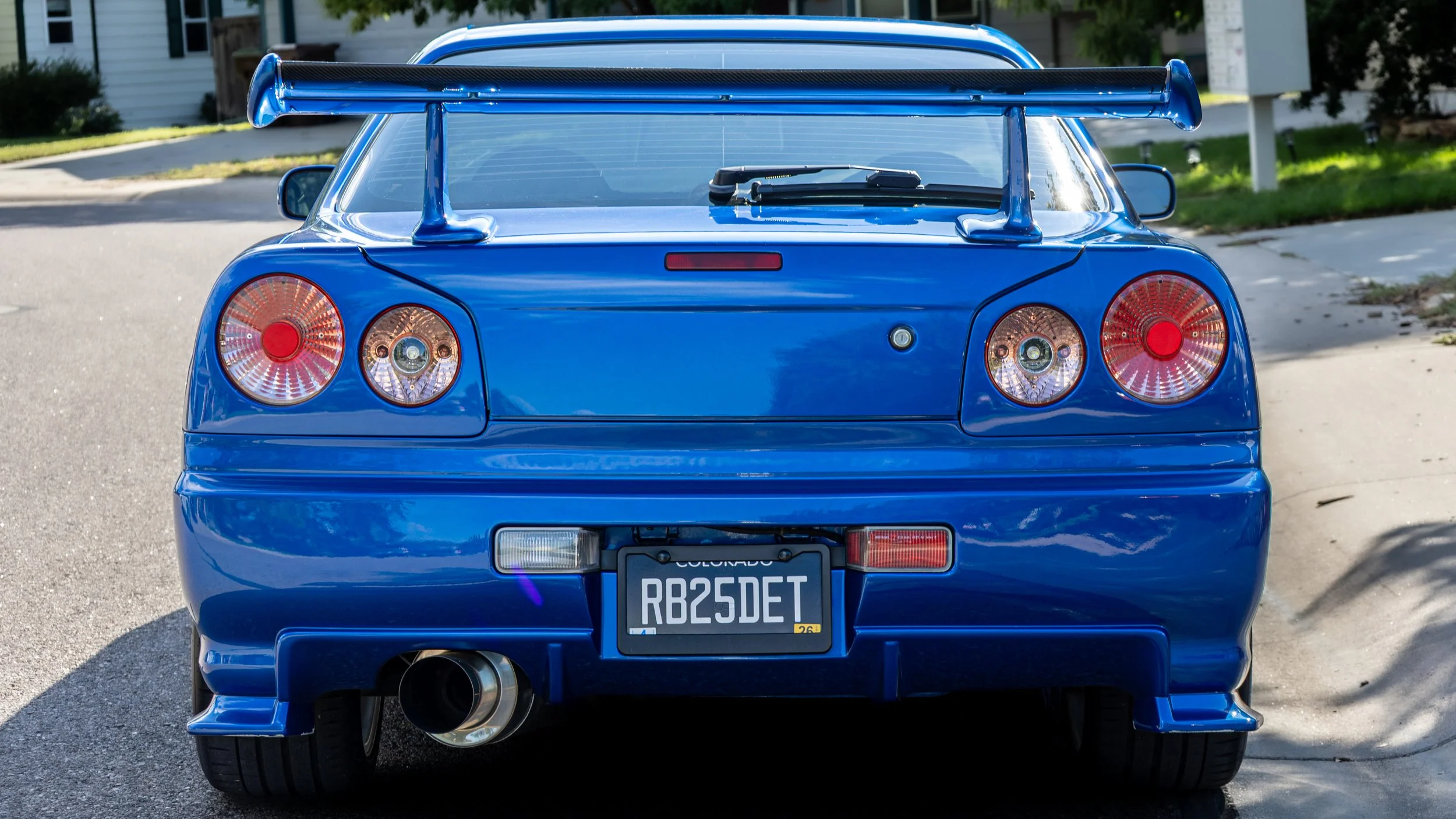 Rear view of a bright blue sports car with a large rear wing, distinctive circular taillights, a Colorado license plate, and a single exhaust pipe in a residential neighborhood.
