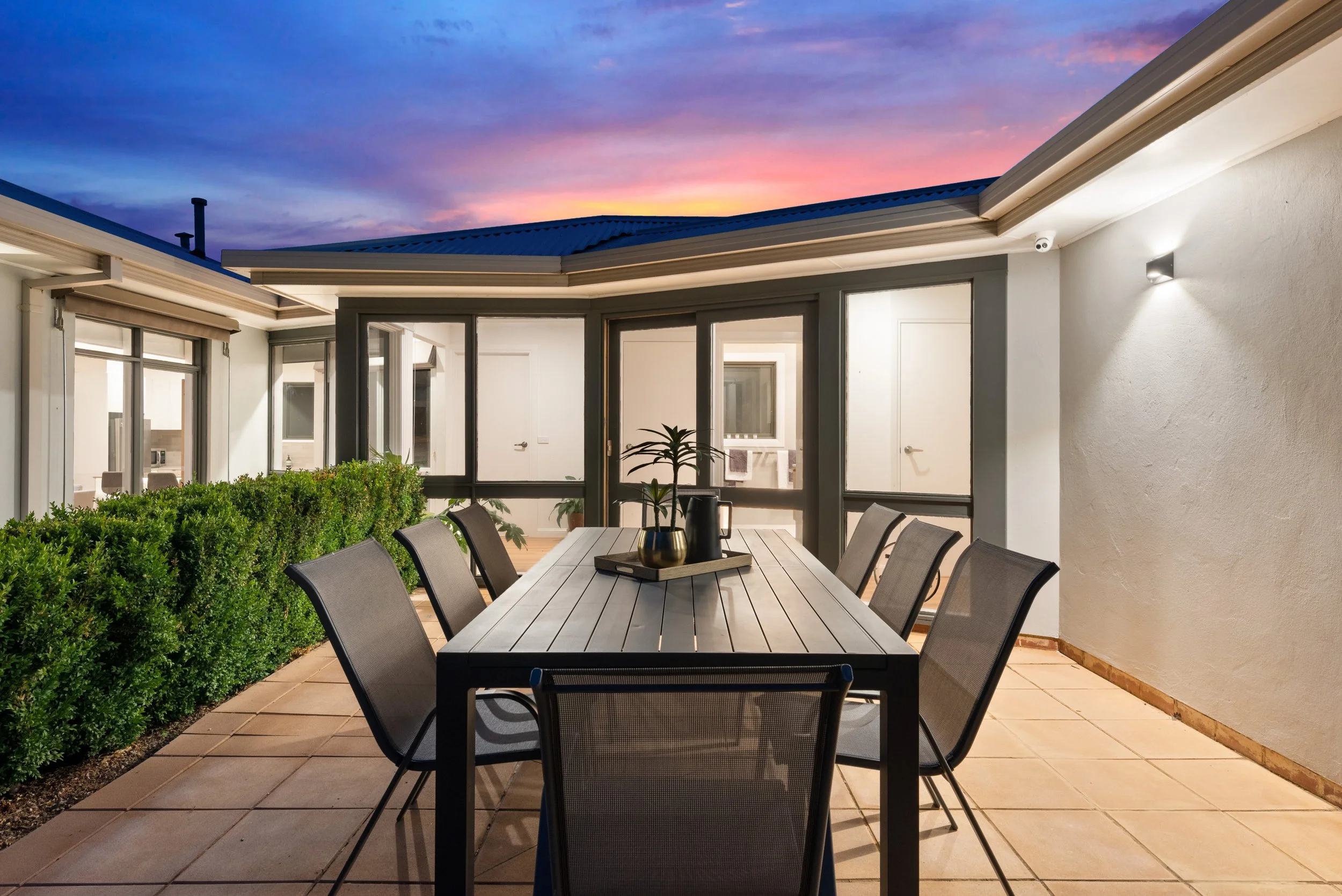 A covered outdoor patio with a large dining table and eight chairs, decorative vases, green bushes along the wall, windows and sliding glass doors, and a sunset sky with purple, pink, and orange clouds.