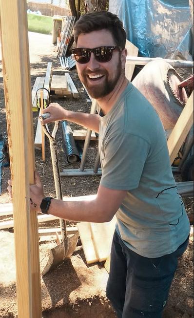 A man with sunglasses smiling and working outdoors on a construction or carpentry project, holding a hammer and standing by a wooden frame.