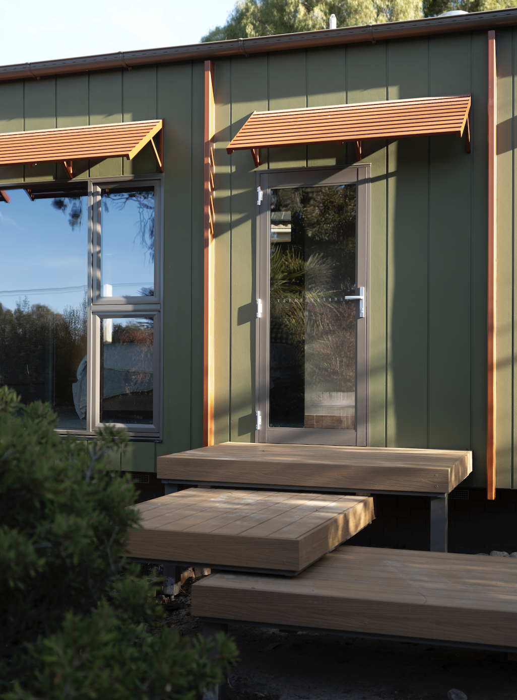 Exterior view of a modern house with green siding, small wooden awnings above windows, and a door with a glass panel. There is a small wooden deck at the entrance.