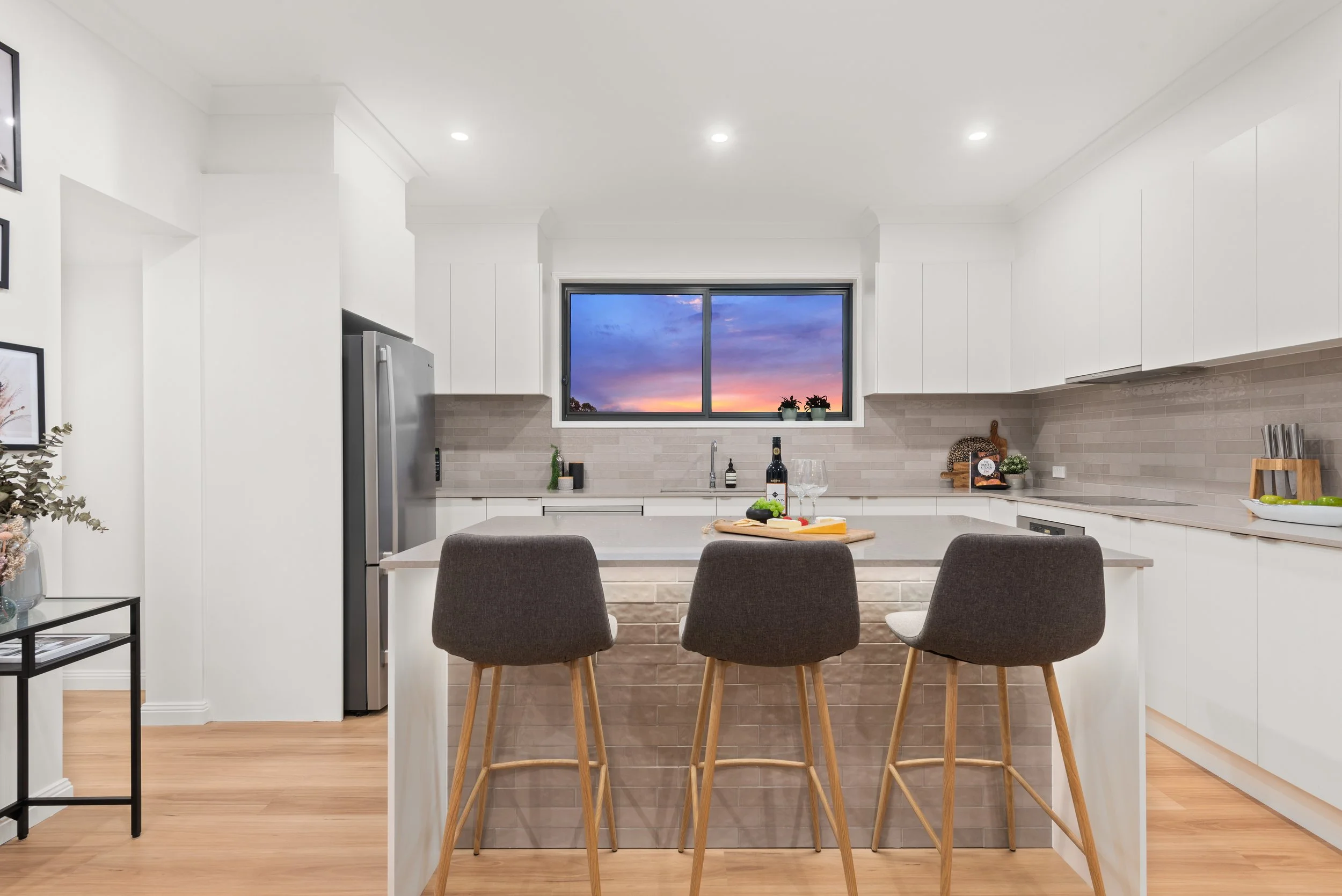 Modern kitchen with white cabinets, gray backsplash, and a large window showing a colorful sunset sky. Kitchen island with three gray chairs, wine bottle, and various decorative items.