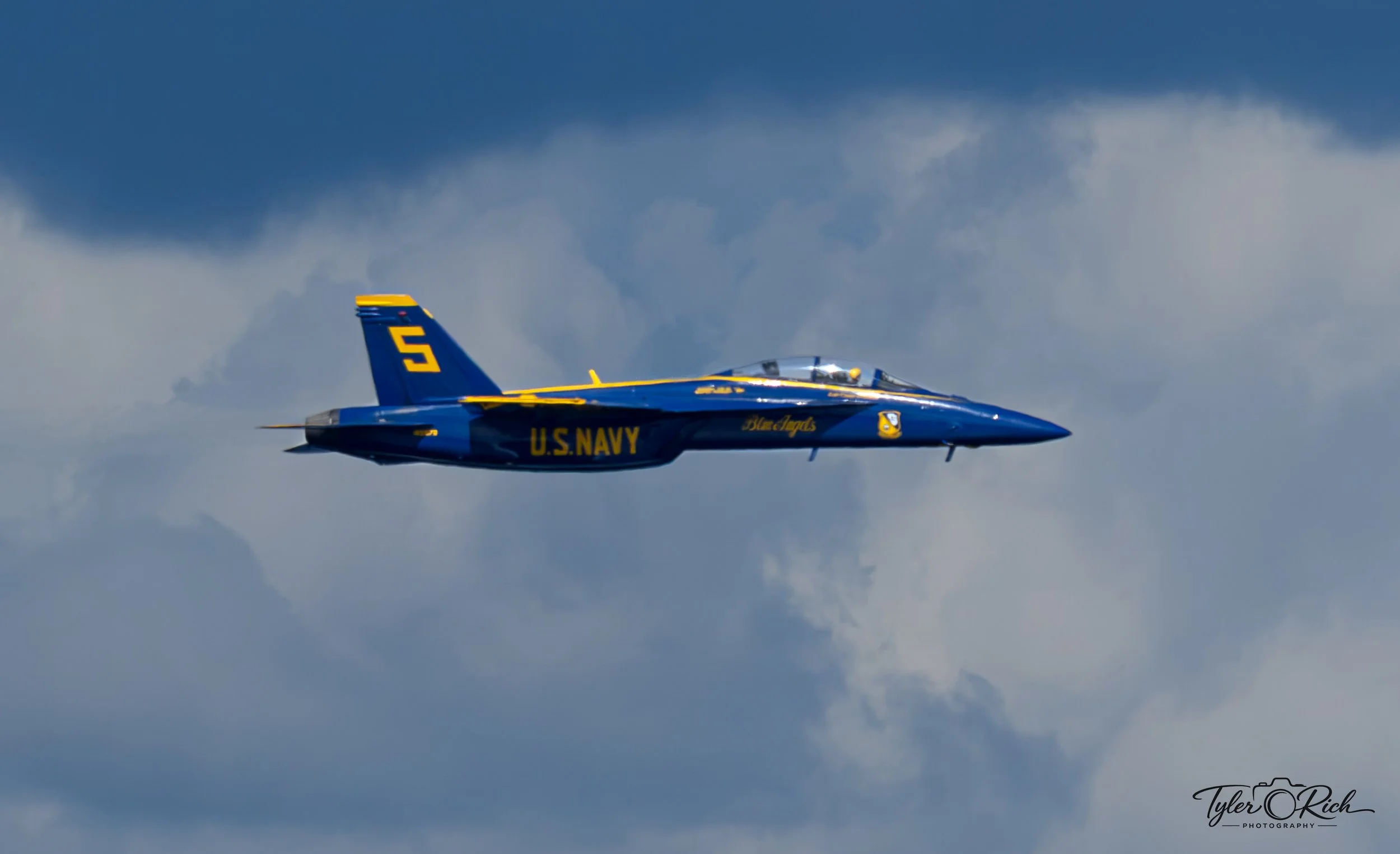 A US Navy Blue Angels fighter jet flying through a partly cloudy sky.