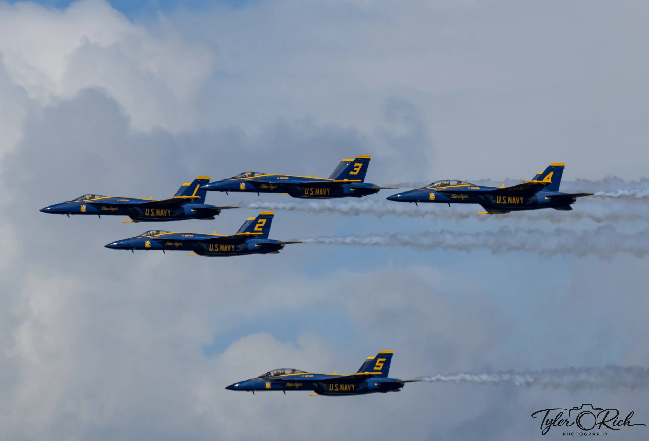 A formation of four U.S. Navy blue and yellow jet aircraft flying in close formation against a partly cloudy sky.