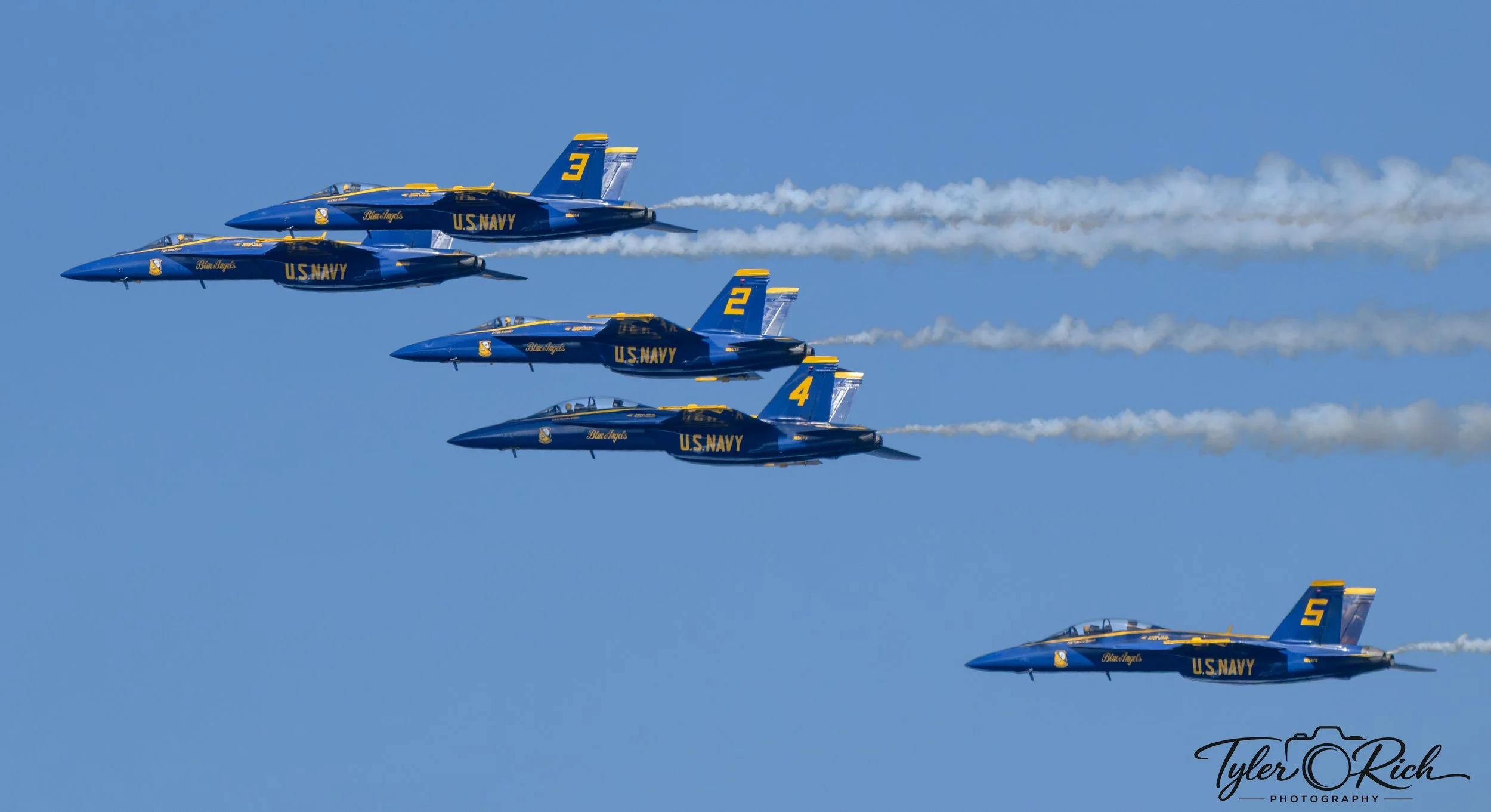 Four U.S. Navy Blue Angels fighter jets flying in a formation against a clear blue sky, with smoke trails behind them during an air show.